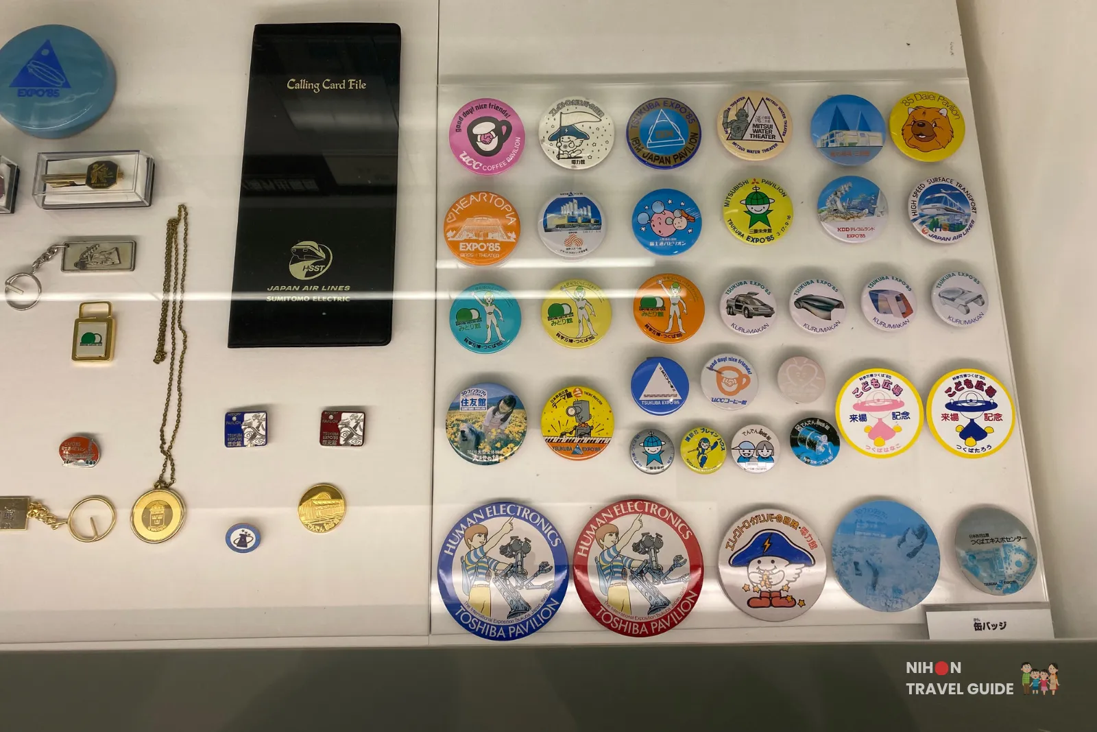 Collection of commemorative badges, pins, keychains, and small souvenirs from the 1985 Tsukuba Expo, including items from different pavilions and companies.