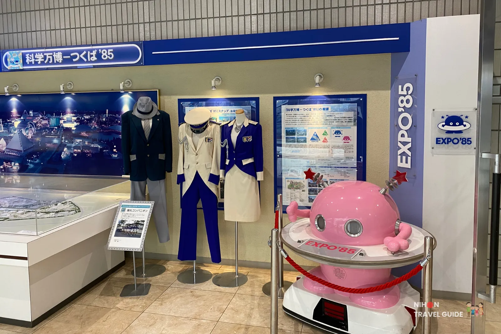Display of official companion (hostess) uniforms and the iconic pink mascot robot Cosmo Hoshimaru in the Tsukuba Expo '85 Memorial. A large screen shows the illuminated Expo site at night.