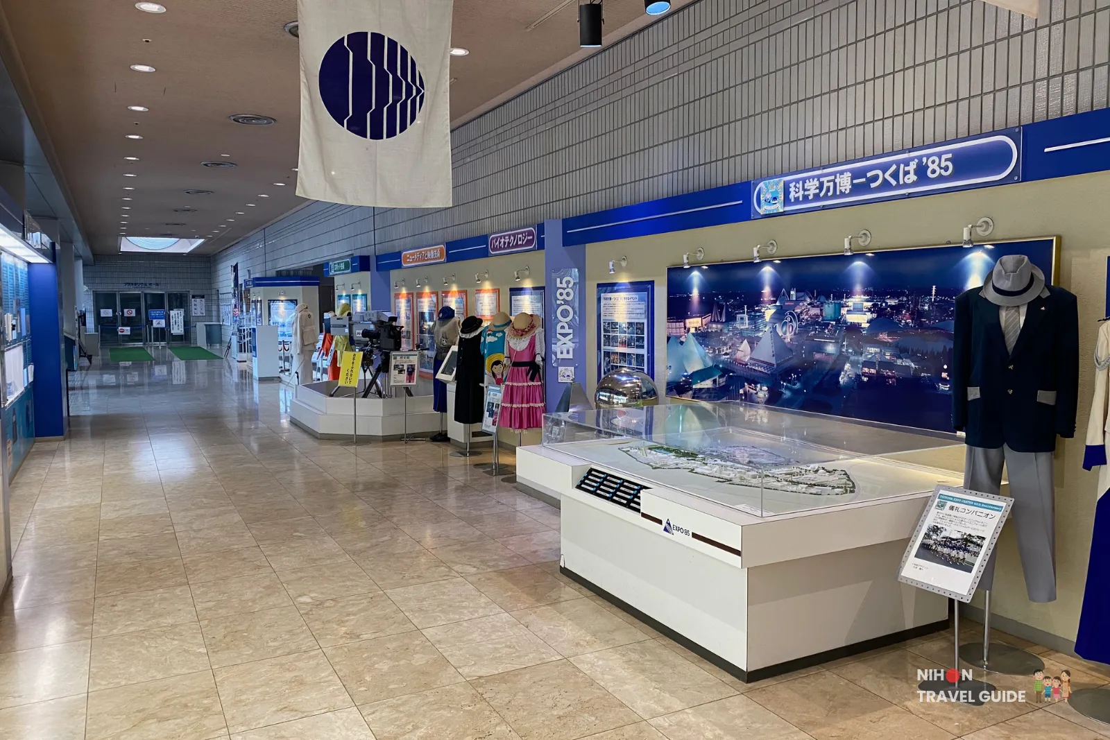 Wide view inside the Tsukuba Expo '85 Memorial exhibition on the first floor. The corridor features large displays, mannequins in companion uniforms, models, and a big night-time photo of the Expo grounds.