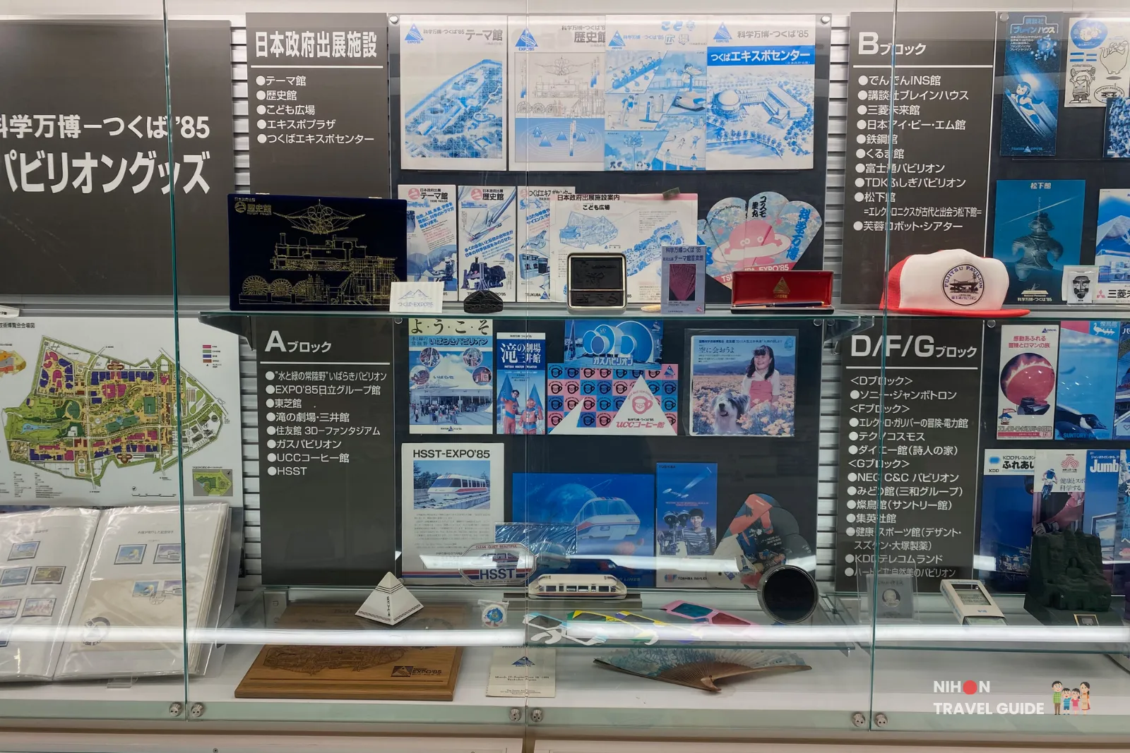 Detailed glass display case in the Tsukuba Expo '85 Memorial showing maps, pamphlets, models, and goods from various pavilions including A Block, B Block, and D/F/G Block of the 1985 Tsukuba Expo.