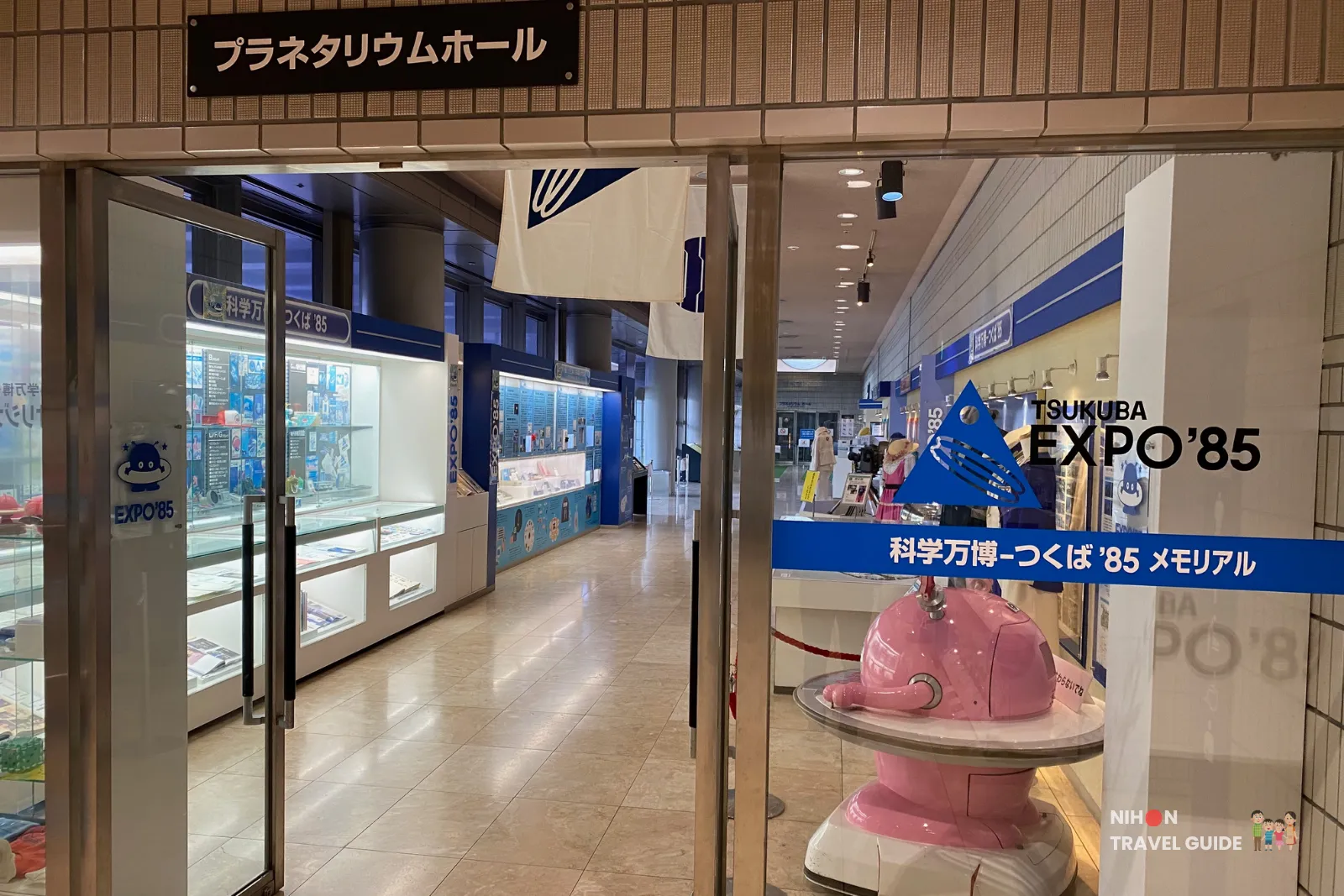 Entrance to the Tsukuba Expo '85 Memorial exhibition and Planetarium Hall on the first floor. The pink mascot robot Cosmo Hoshimaru welcomes visitors, with display cases full of 1985 Expo memorabilia visible inside.