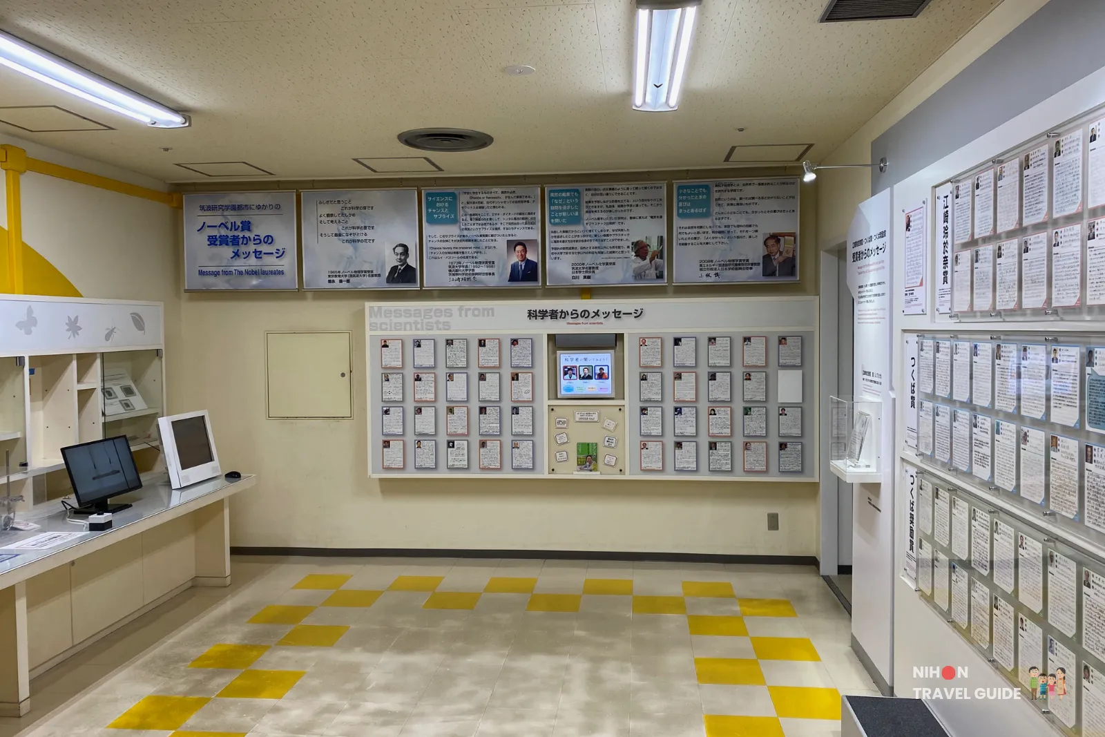 Museum exhibit wall with many small plaques and photos under a sign 'Messages from scientists' in English and Japanese; computer desks to the left; yellow checkered floor tiles.