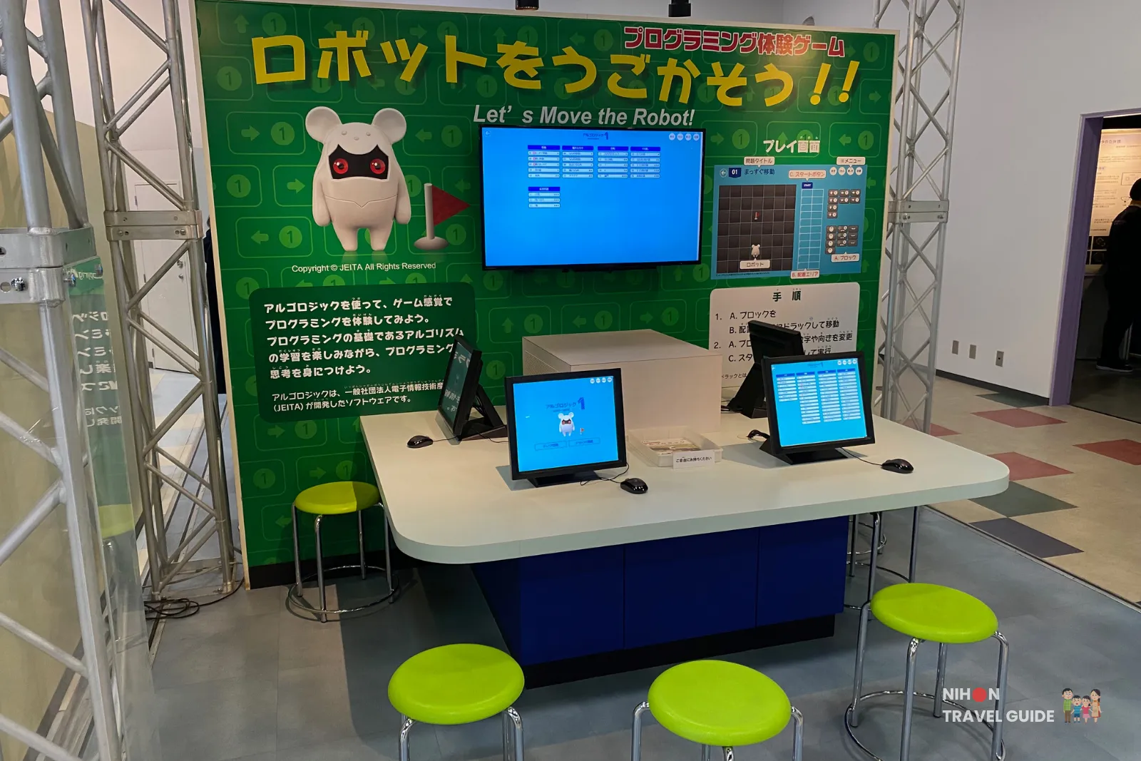Interactive programming booth featuring a white robot mascot on a bright green backdrop, a wall monitor, and tablets on a curved white counter with lime stools nearby