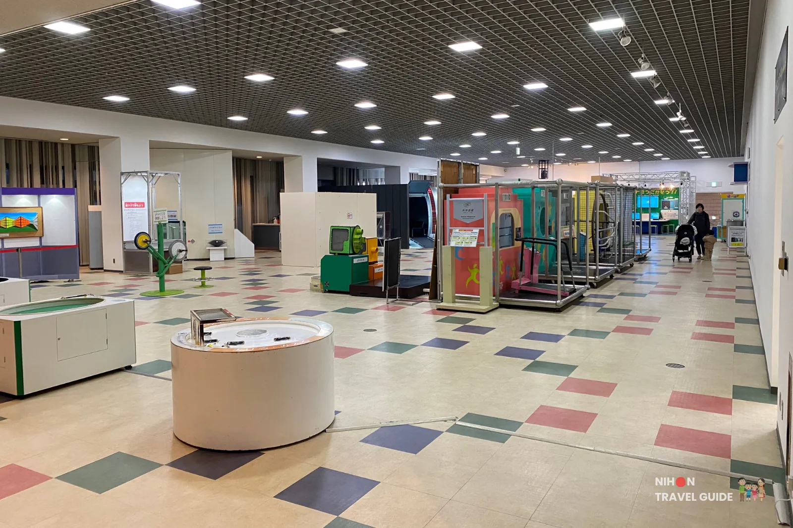 Wide interior view of the Fun Science Zone on the first floor of Tsukuba Expo Center. The large open hall features many interactive science exhibits, colorful flooring, and various hands-on stations under a high grid ceiling.