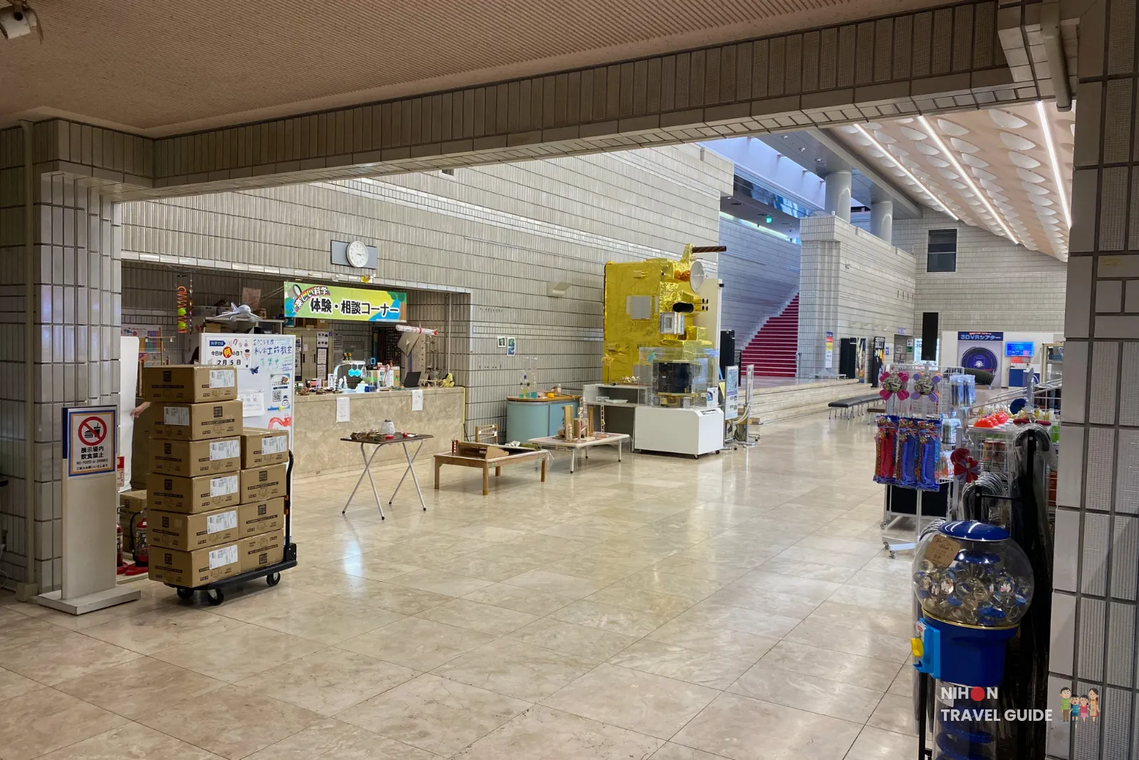 View of the Science Museum Shop and experience/consultation corner at Tsukuba Expo Center, with shelves of merchandise, a large yellow satellite model, and stacked boxes in the foreground.