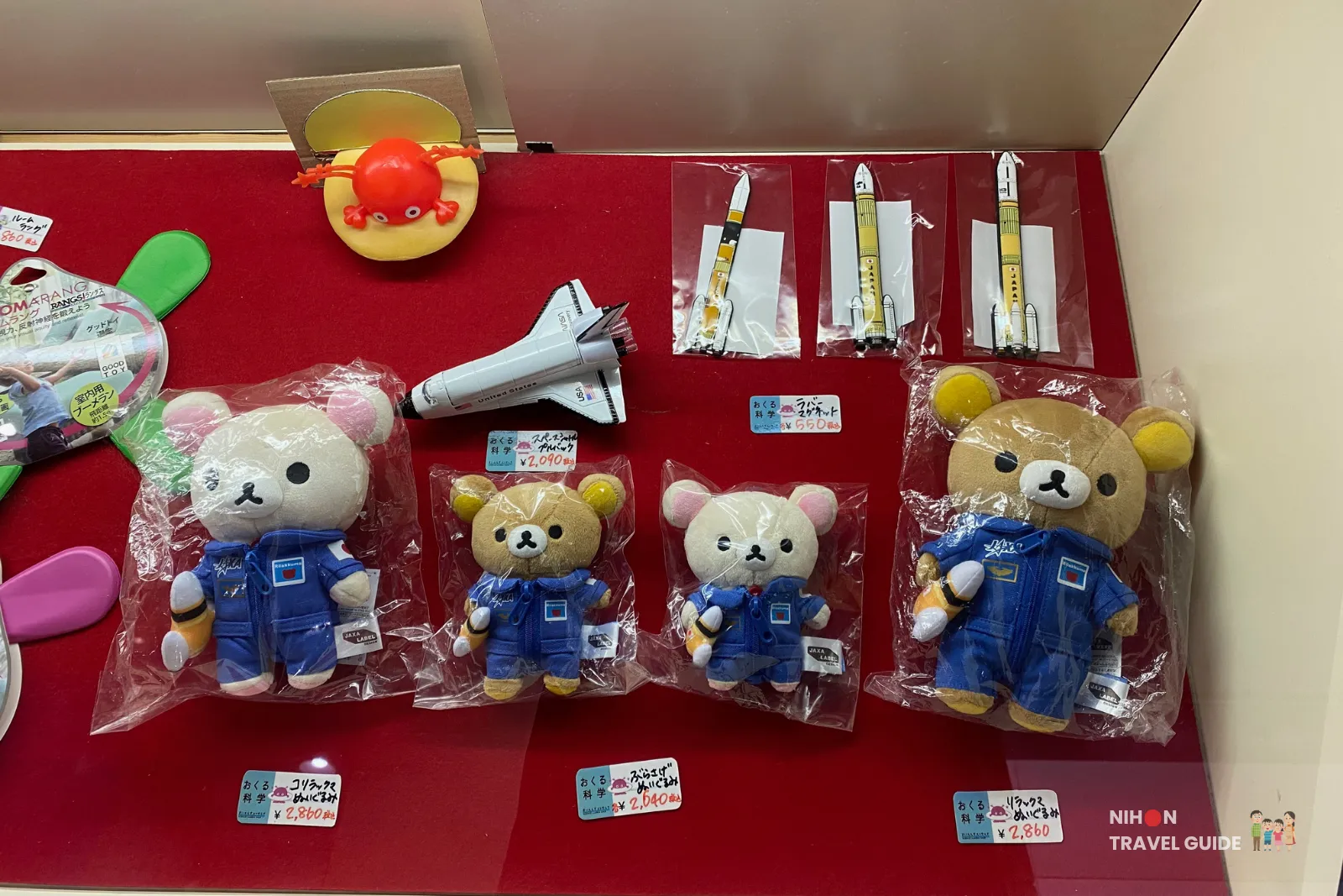 Gift shop display at Tsukuba Expo Center featuring Rilakkuma plush toys dressed in JAXA astronaut suits, along with space shuttle and rocket models. Popular science-themed souvenirs for visitors.