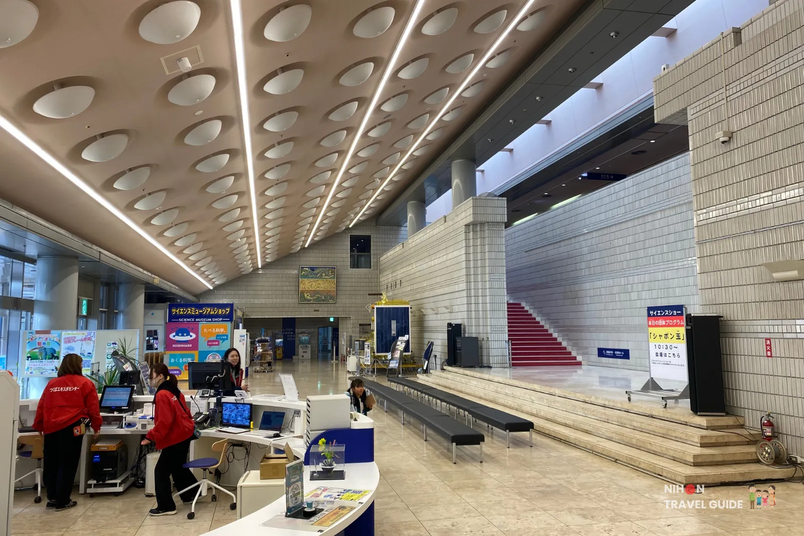 Spacious main lobby and information counter inside Tsukuba Expo Center. Staff in red jackets assist visitors at the reception desk, with the high ceiling, planetarium shop, and grand staircase visible in the background.