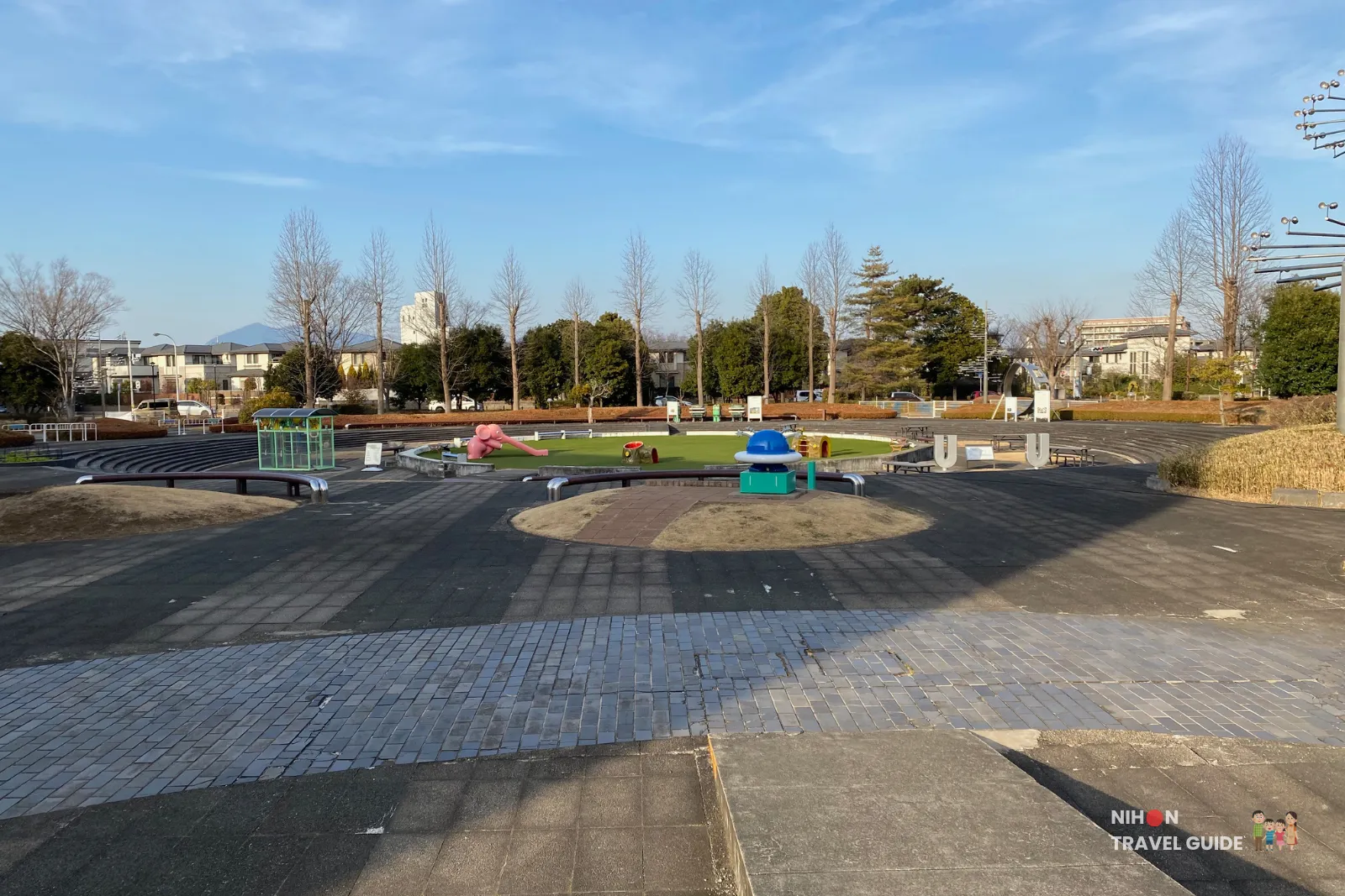 Outdoor exhibition and playground area at Tsukuba Expo Center. Features a circular grassy amphitheater with play equipment, picnic tables, and modern sculptures under a clear blue sky.