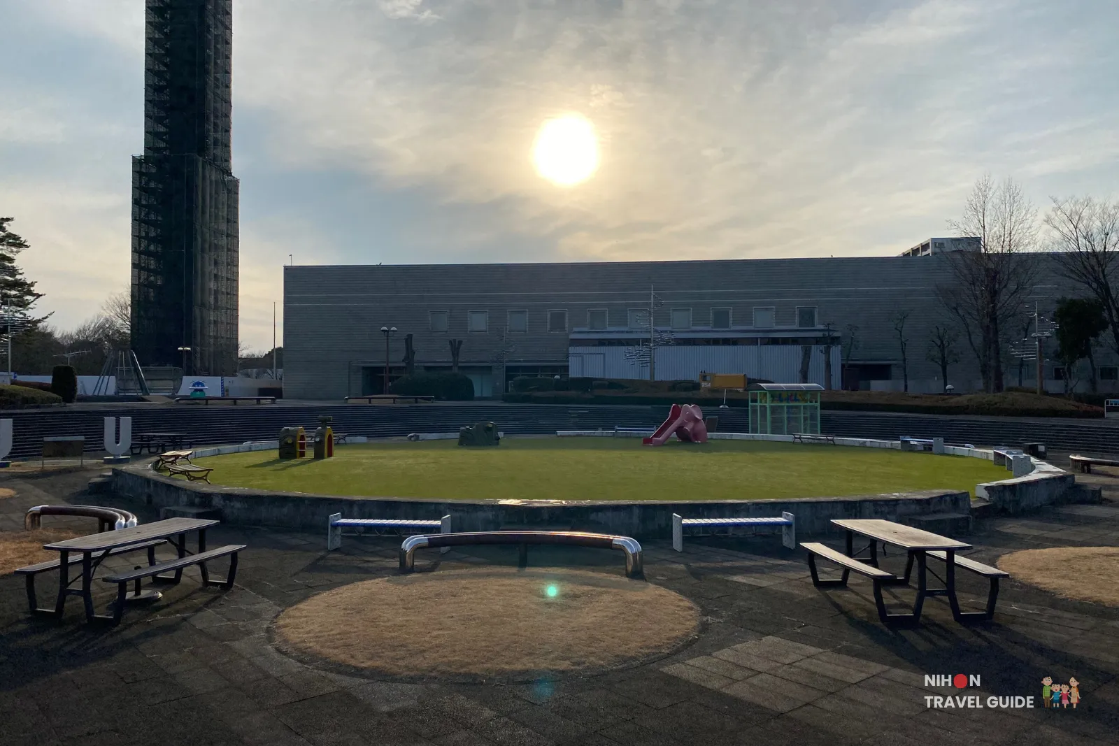 Family-friendly outdoor area at Tsukuba Expo Center with playground equipment, picnic benches, and the tall H-II rocket model visible in the background on a sunny day.
