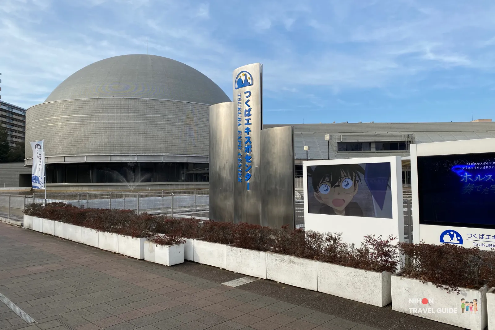 Large gray dome-shaped science center with a tall metallic sign and a screen displaying a surprised anime character.