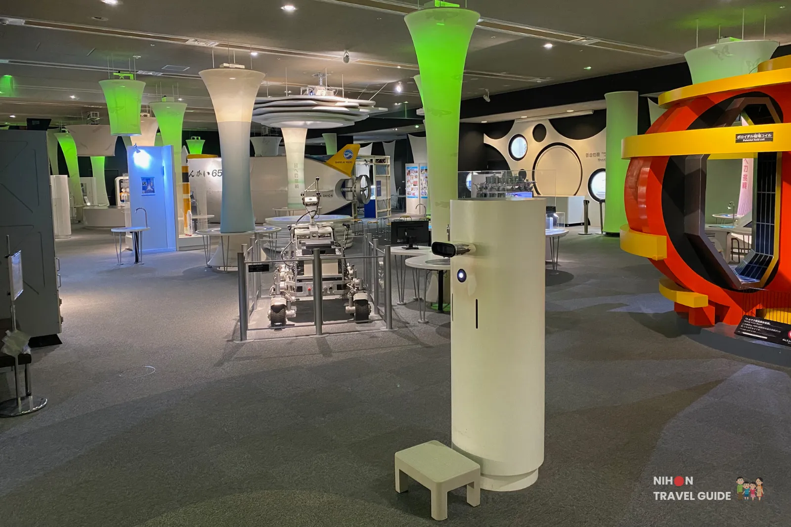 Wide view of the second floor exhibition area at Tsukuba Expo Center. The spacious hall features futuristic green and white towers, interactive stations, a robot display, and colorful modern design elements.