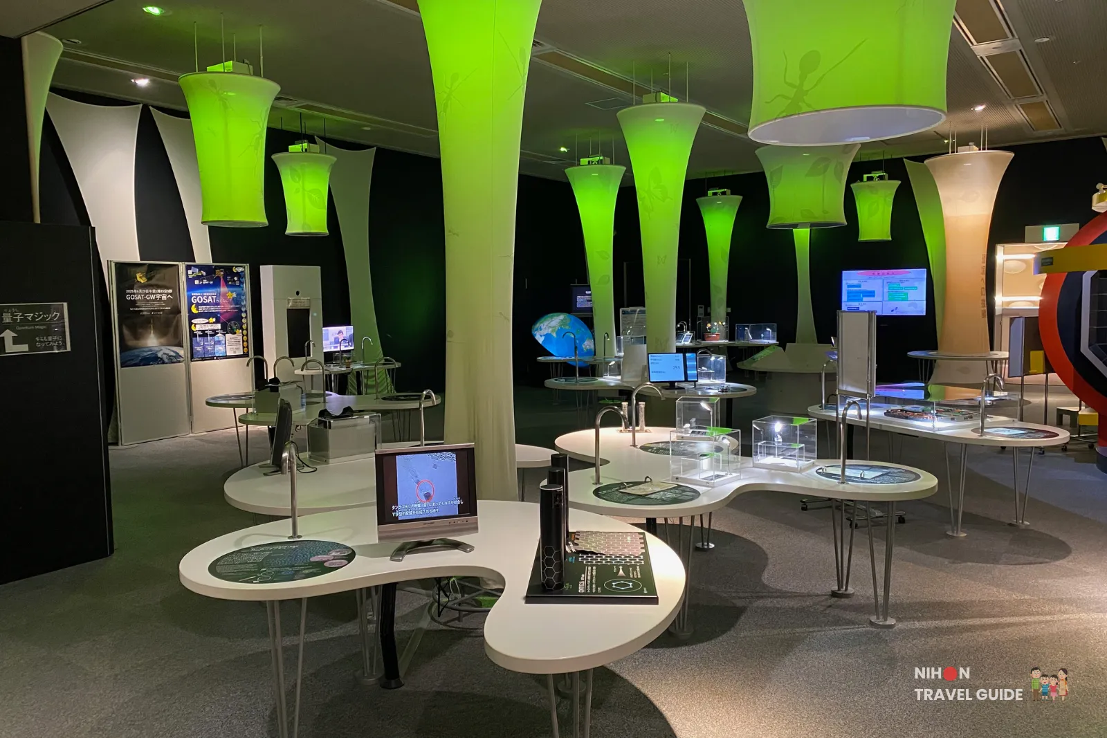 Interactive science stations on the second floor of Tsukuba Expo Center, illuminated by large glowing green pendant lights. The area includes touch-screen displays and hands-on exhibits focused on advanced science topics.