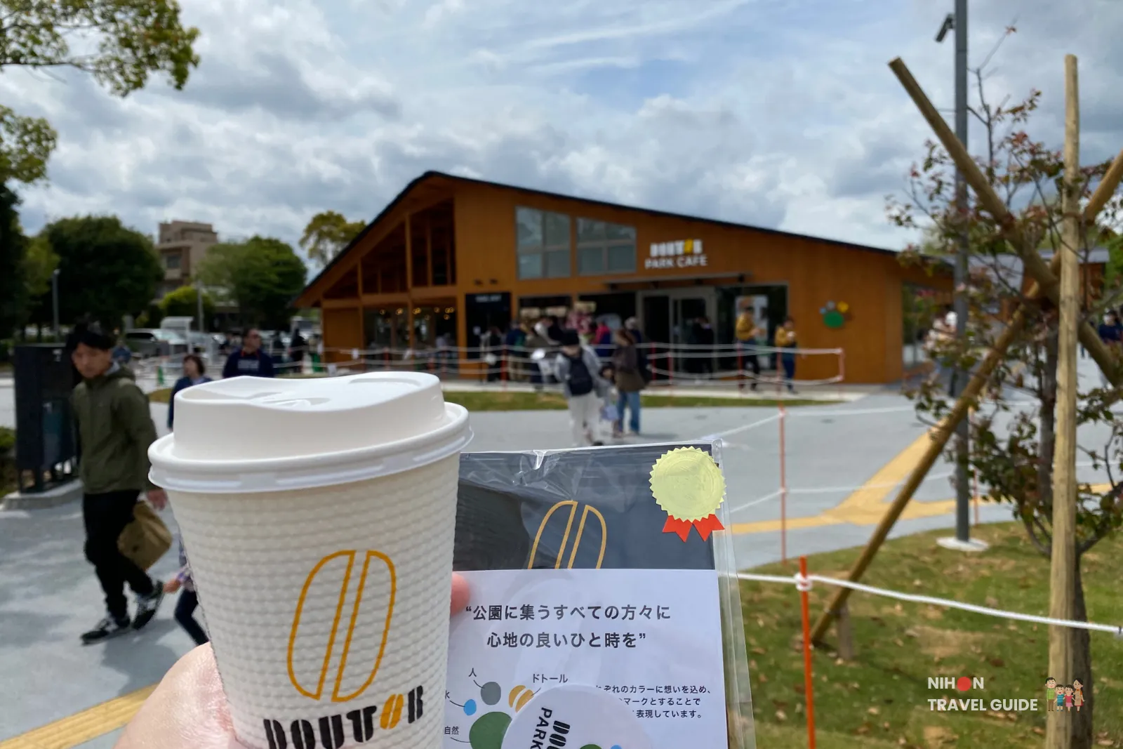 doutor-park-cafe-coffee-gift-mito-kobun-terrace Hand holding a Doutor coffee cup and complimentary gift (drip coffee pack + sticker) at Doutor Park Cafe (ドトール パークカフェ) in Mito Kobun Terrace. The gift features the shop concept: “公園に集うすべての方々に 心地の良いひと時を” (A pleasant moment for everyone gathering in the park).