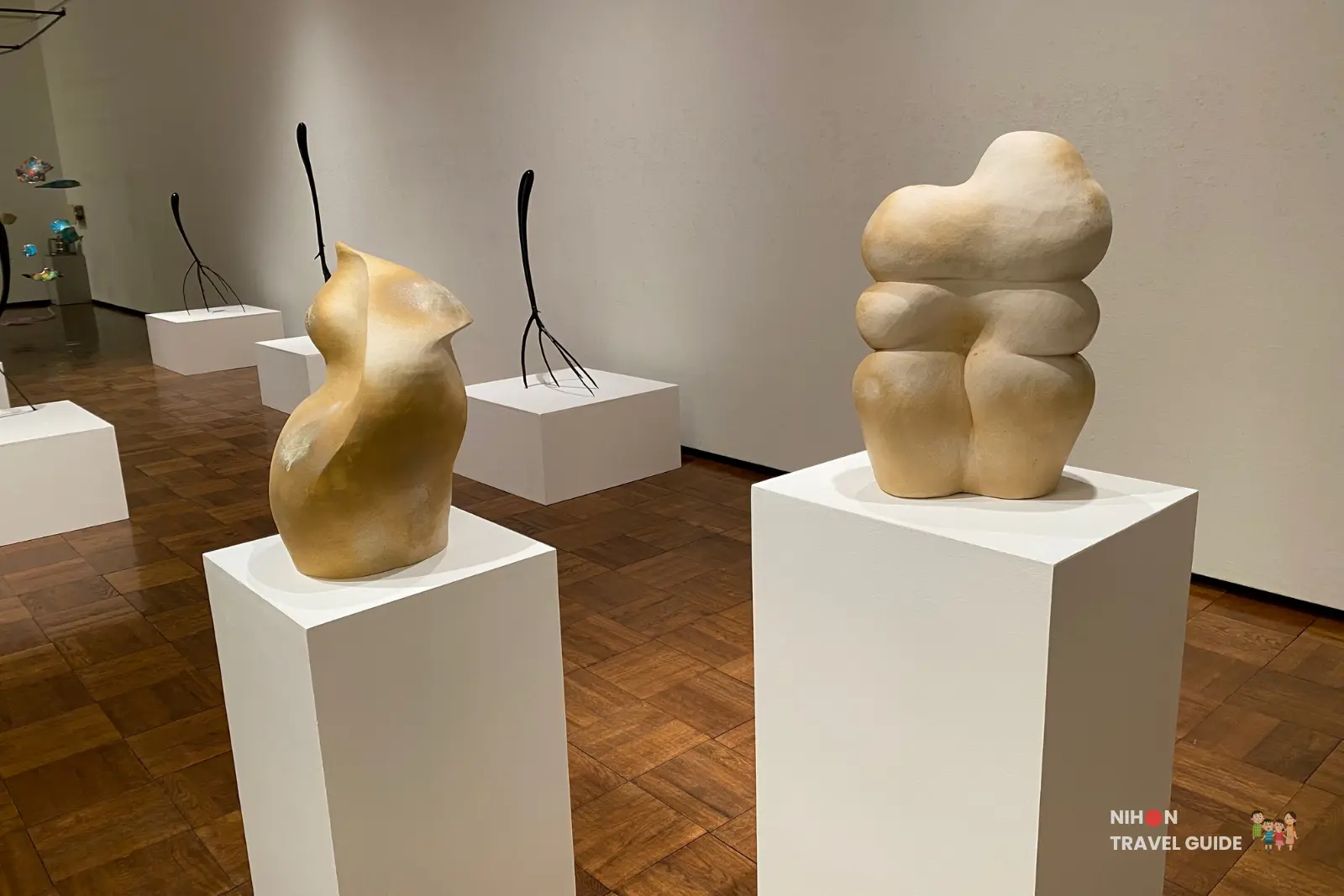 Sculpture gallery detail showing Element-Relation (stacked beige organic forms) and a golden abstract sculpture on white pedestals, with black wire pieces in the background.