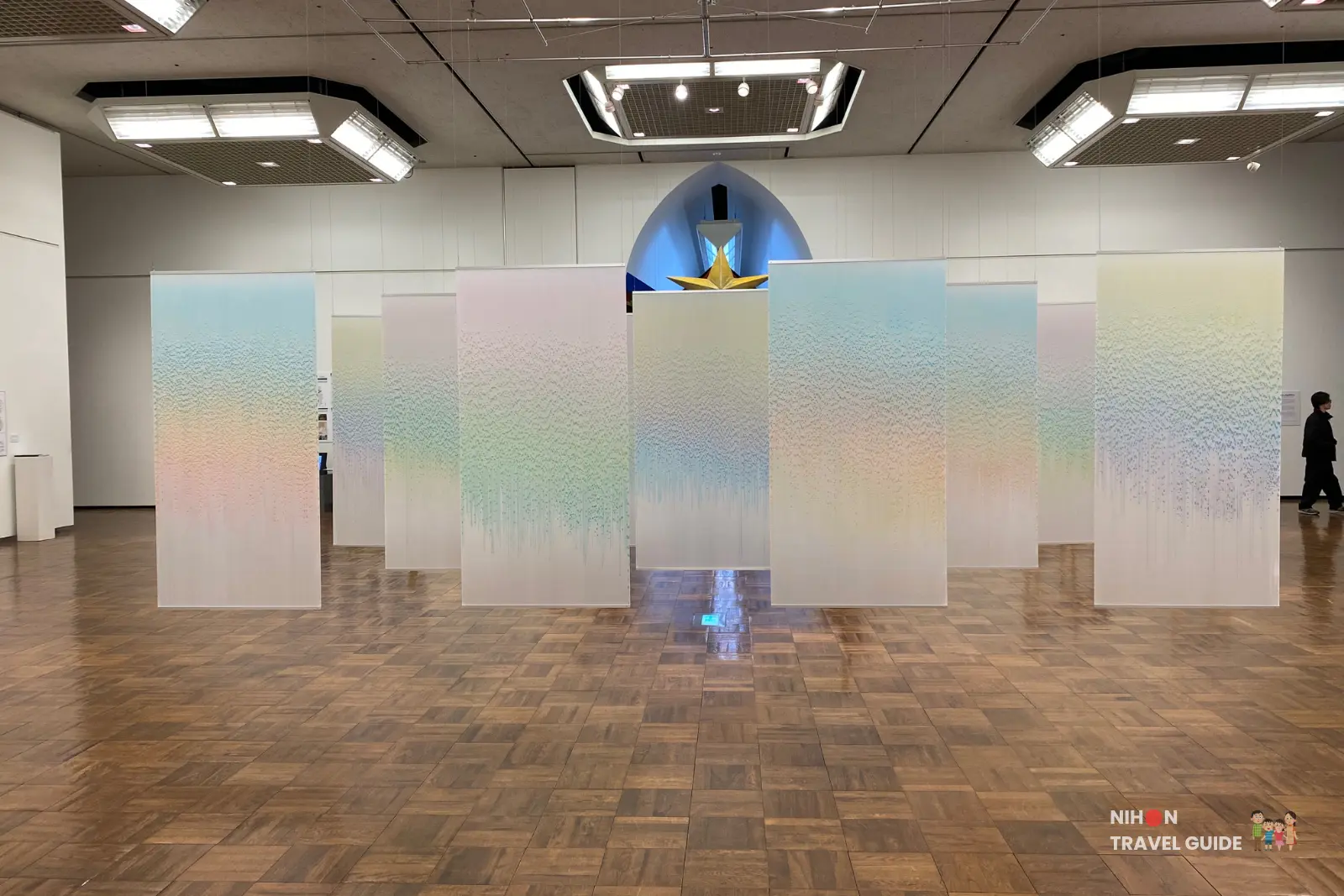 Wide view of Flow installation by Saya Takama — tall translucent gradient panels with pastel colors hanging in an arc across the main gallery, golden star sculpture visible in the background alcove.