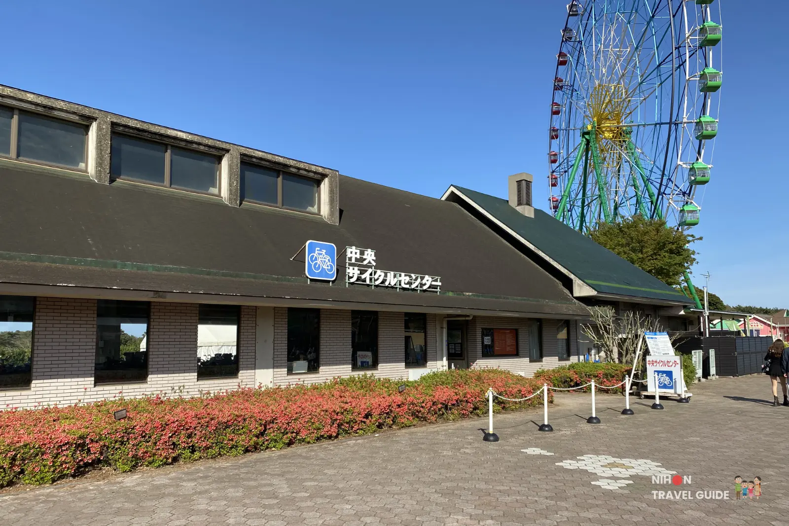 Central Cycle Center building at Hitachi Seaside Park where visitors can rent bicycles. The iconic Blue Eyes Ferris Wheel of the Pleasure Garden stands tall in the background on a clear sunny day.