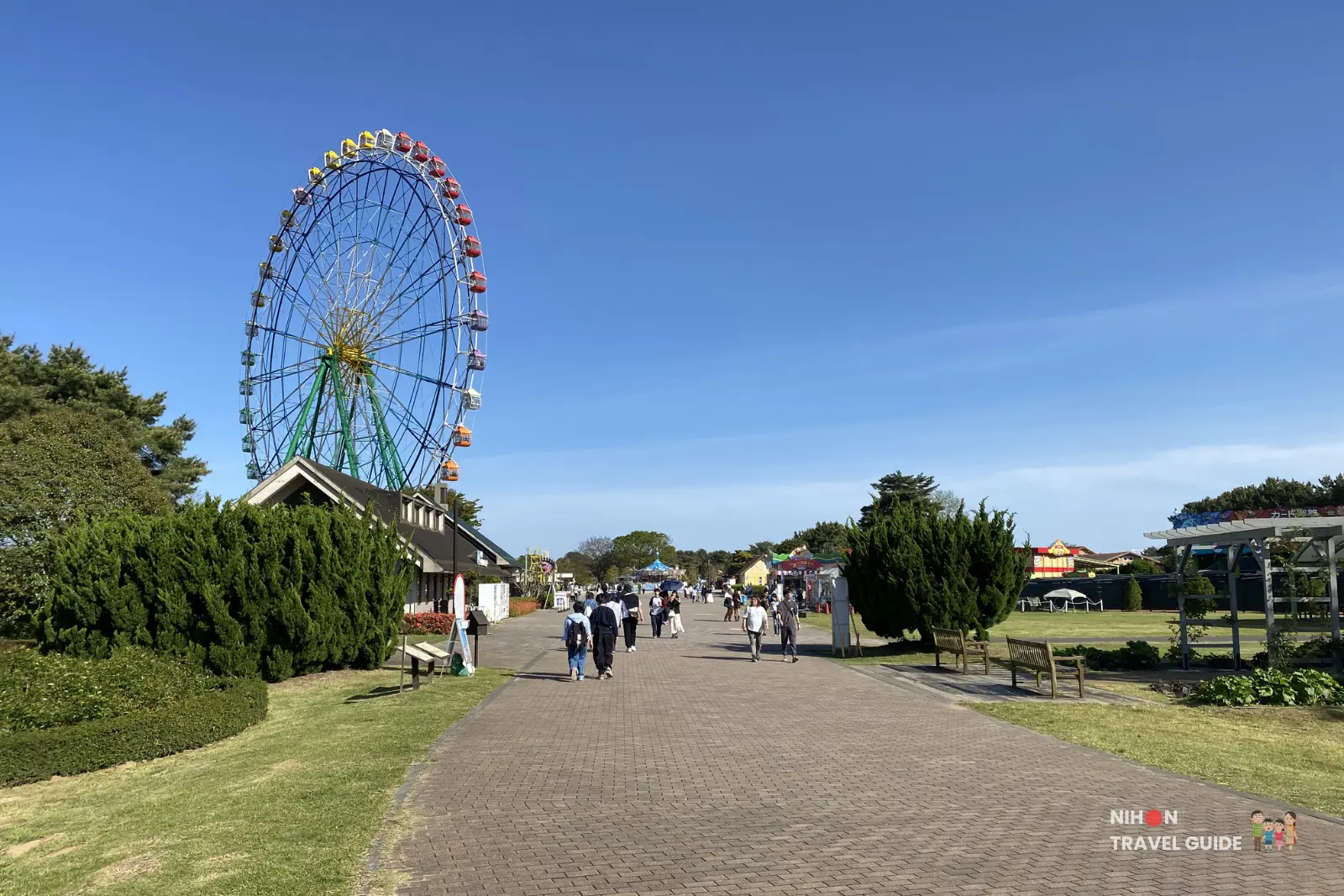 View of the Pleasure Garden (amusement park zone) at Hitachi Seaside Park. The iconic Blue Eyes Ferris Wheel stands tall against the blue sky, with a wide brick path leading toward rides and attractions.