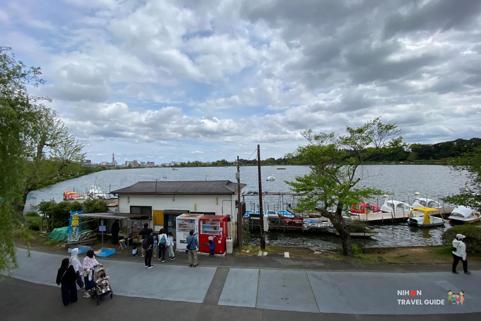 Overview of Ishikawa Boat Rental Shop area at Lake Senba in Mito, showing the rental building, visitors, vending machines, and swan boats on the water with the lake and city skyline in the background.
