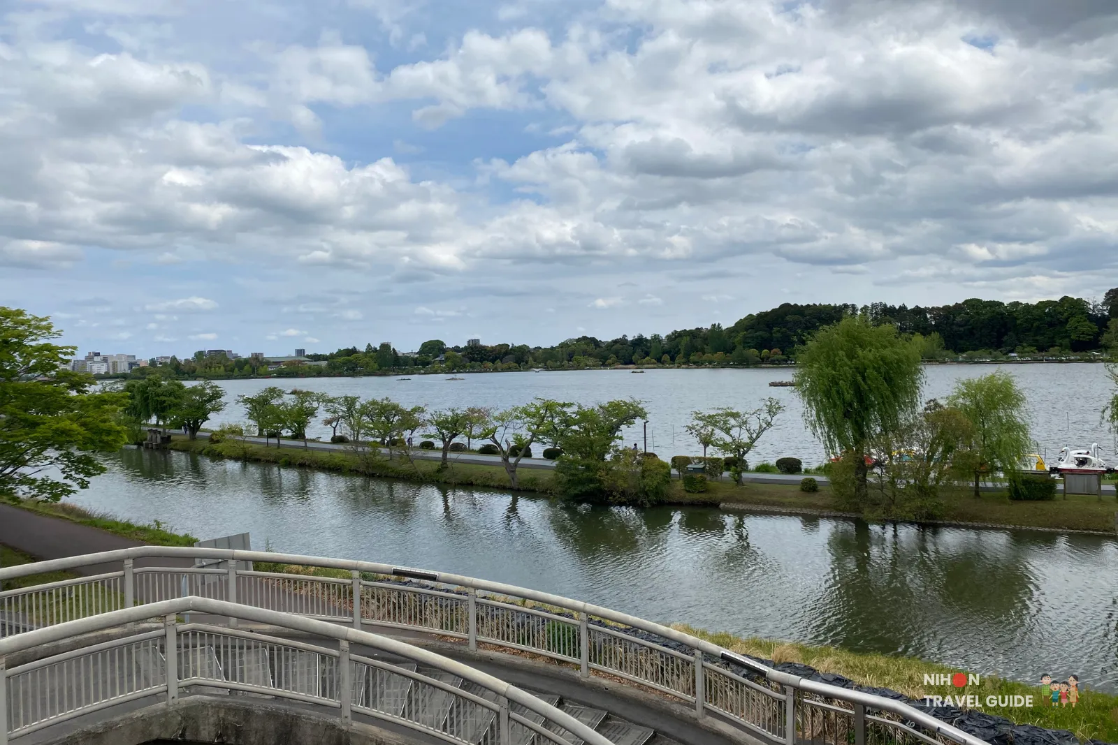 View along the curved pedestrian bridge over Lake Senba toward Kairakuen Garden, showing visitors walking with the lake and waterfront scenery on both sides.