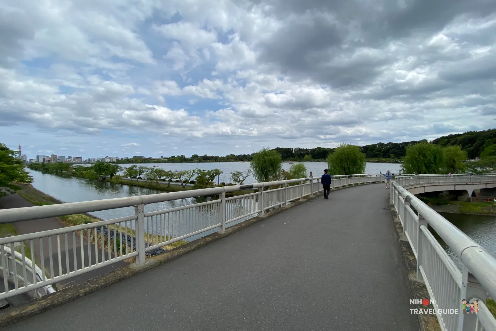 Panoramic view of Lake Senba from the bridge leading toward Kairakuen Garden, with the expansive lake, walking paths, and Mito city skyline under a cloudy sky.