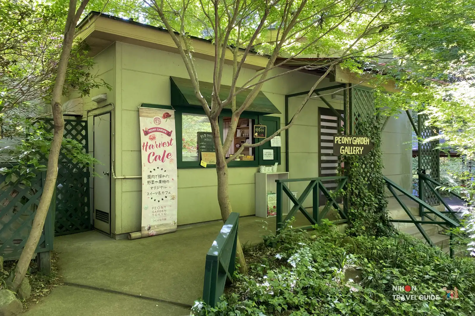 Harvest Cafe and Peony Garden Gallery building at Peony Garden Tokyo, surrounded by lush green trees and dappled sunlight on a bright spring day, with visitors nearby.