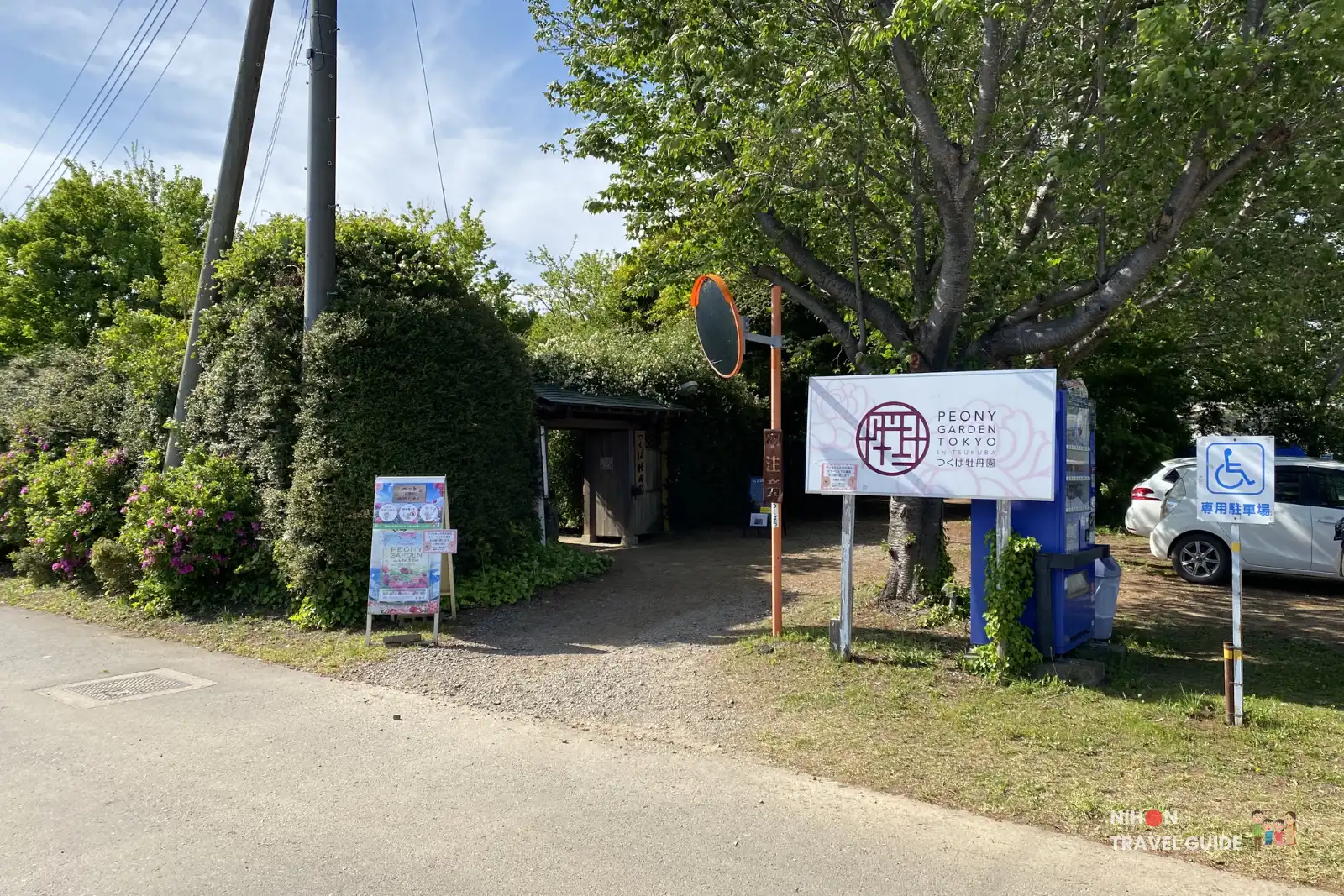 peony-garden-tokyo-main-entrance-gate-sign Main entrance of Peony Garden Tokyo in Tsukuba, featuring the large white garden sign with logo, a blue vending machine, accessibility parking sign, and the path leading into the garden surrounded by greenery on a sunny day.