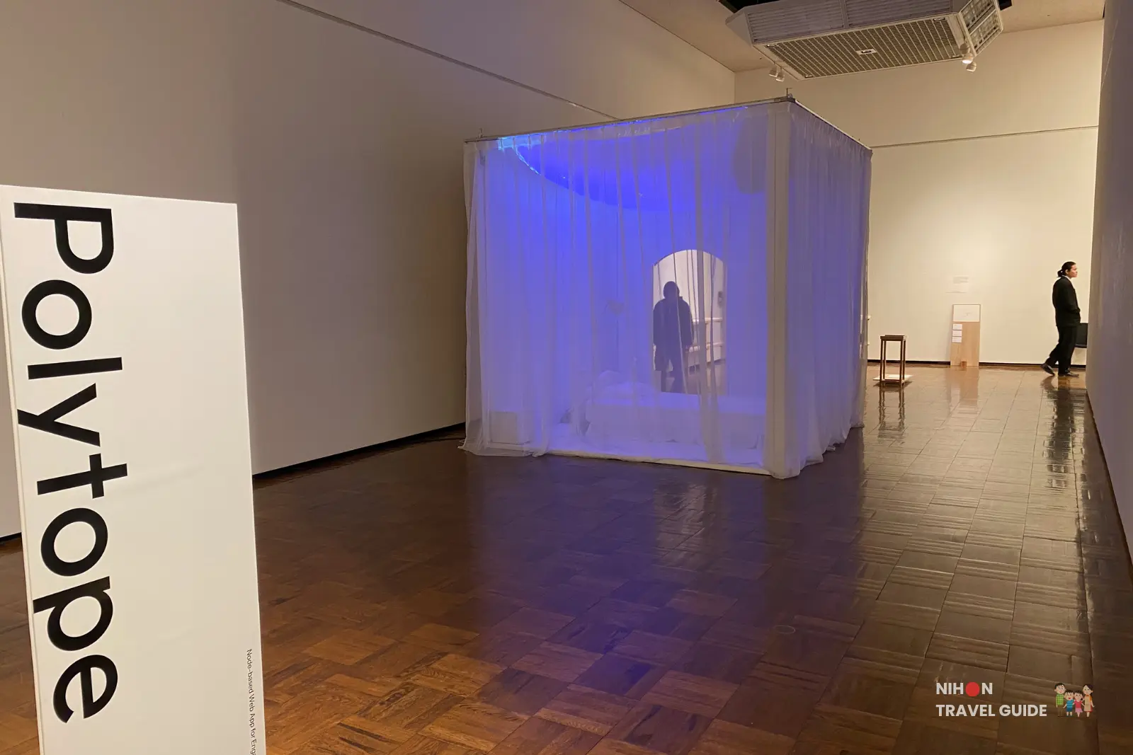 Polytape curtain installation by Taisuke Naganawa — glowing blue translucent cube with bed and silhouette inside, viewed through the white “Polytape” title panel