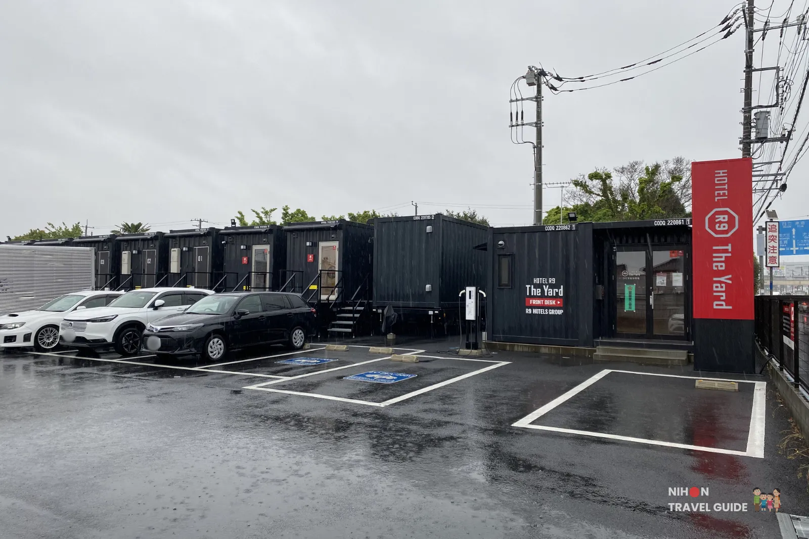 Exterior view of R9 The Yard Hotel Hitachinaka Ichige showing the black container-style buildings and large parking lot on a rainy day in Ibaraki, Japan