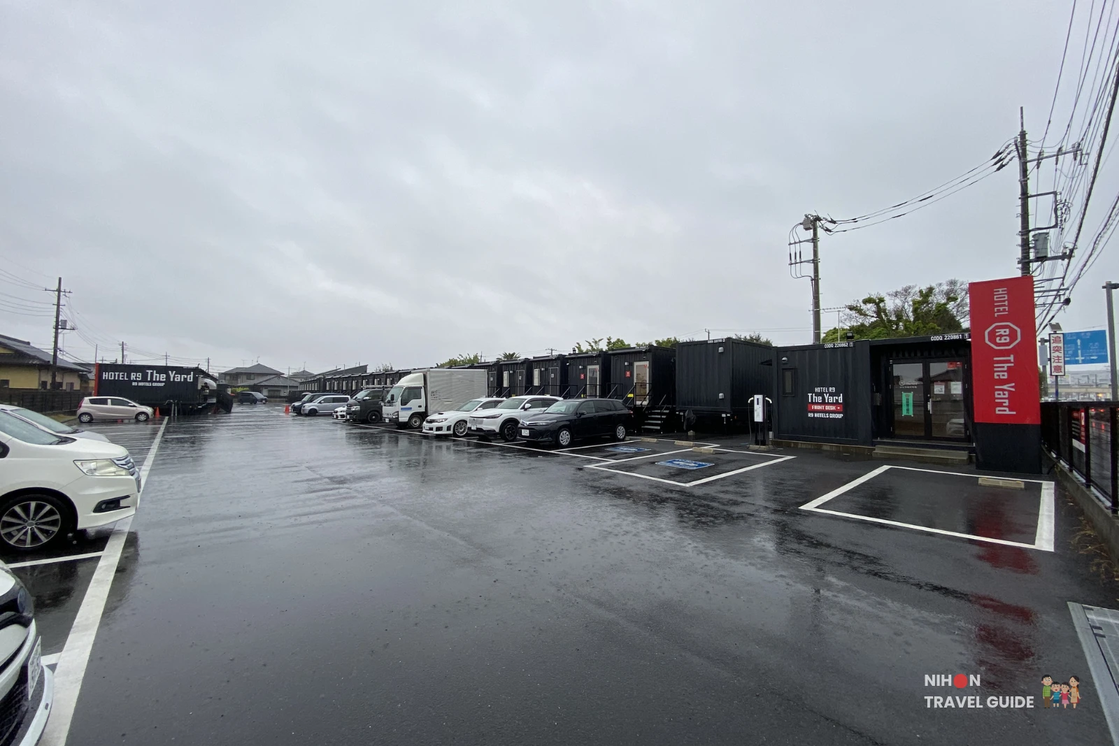 Exterior view of R9 The Yard Hotel Hitachinaka Ichige showing the black container-style buildings and large parking lot on a rainy day in Ibaraki, Japan