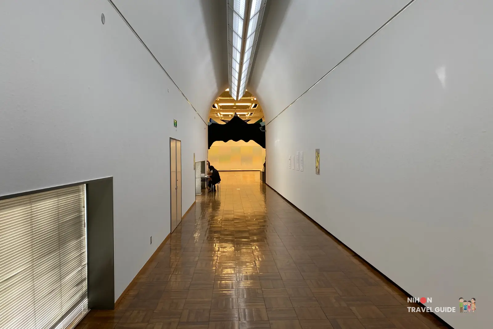 Long white corridor inside Tsukuba Museum of Art leading toward an arched exhibition alcove with a black wavy hanging installation and glowing yellow panels at the far end