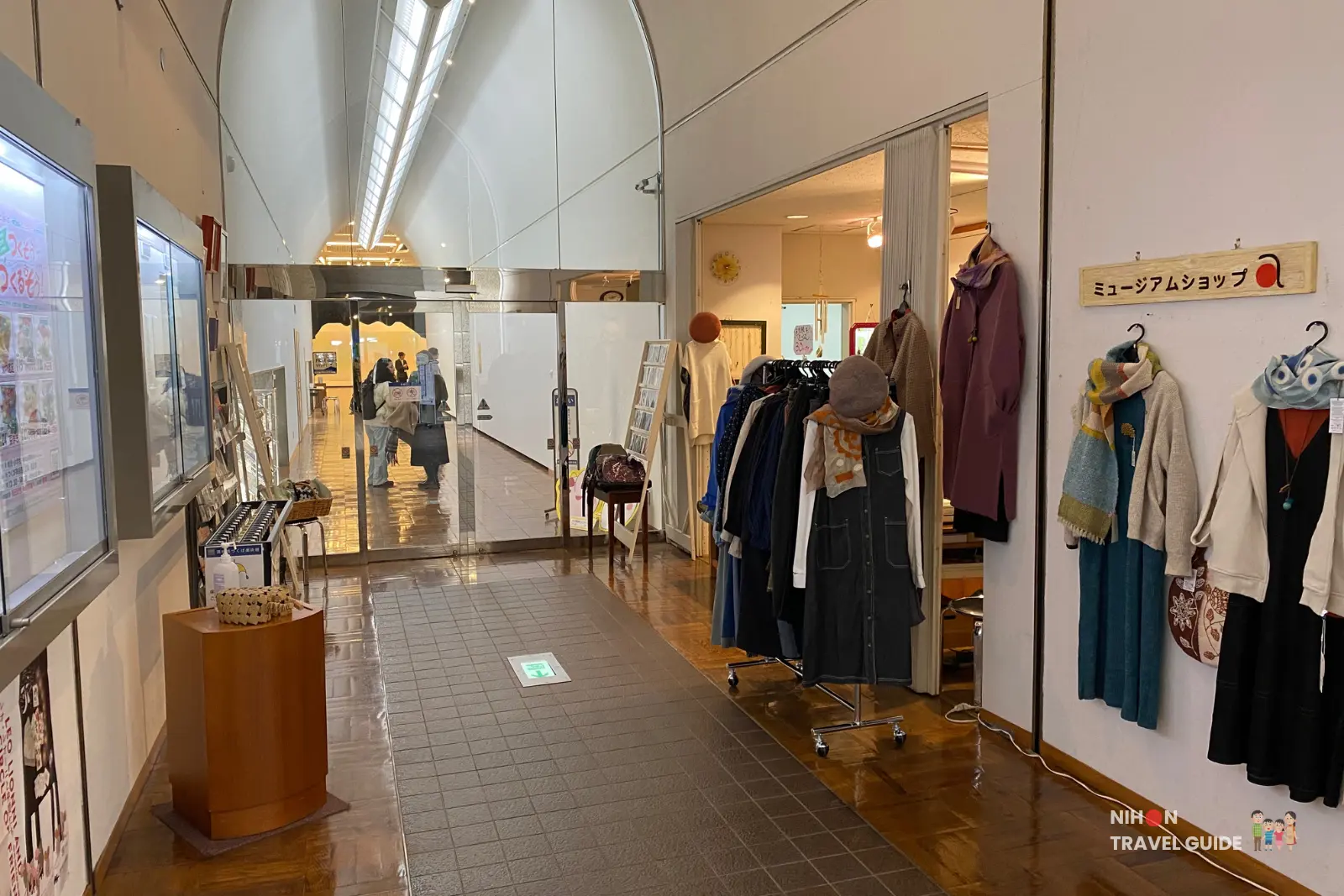 Museum shop at Tsukuba Museum of Art (inside Tsukuba Cultural Hall “Ars”) featuring clothing racks with coats, scarves, and accessories, with the arched hallway leading toward the exhibition galleries.