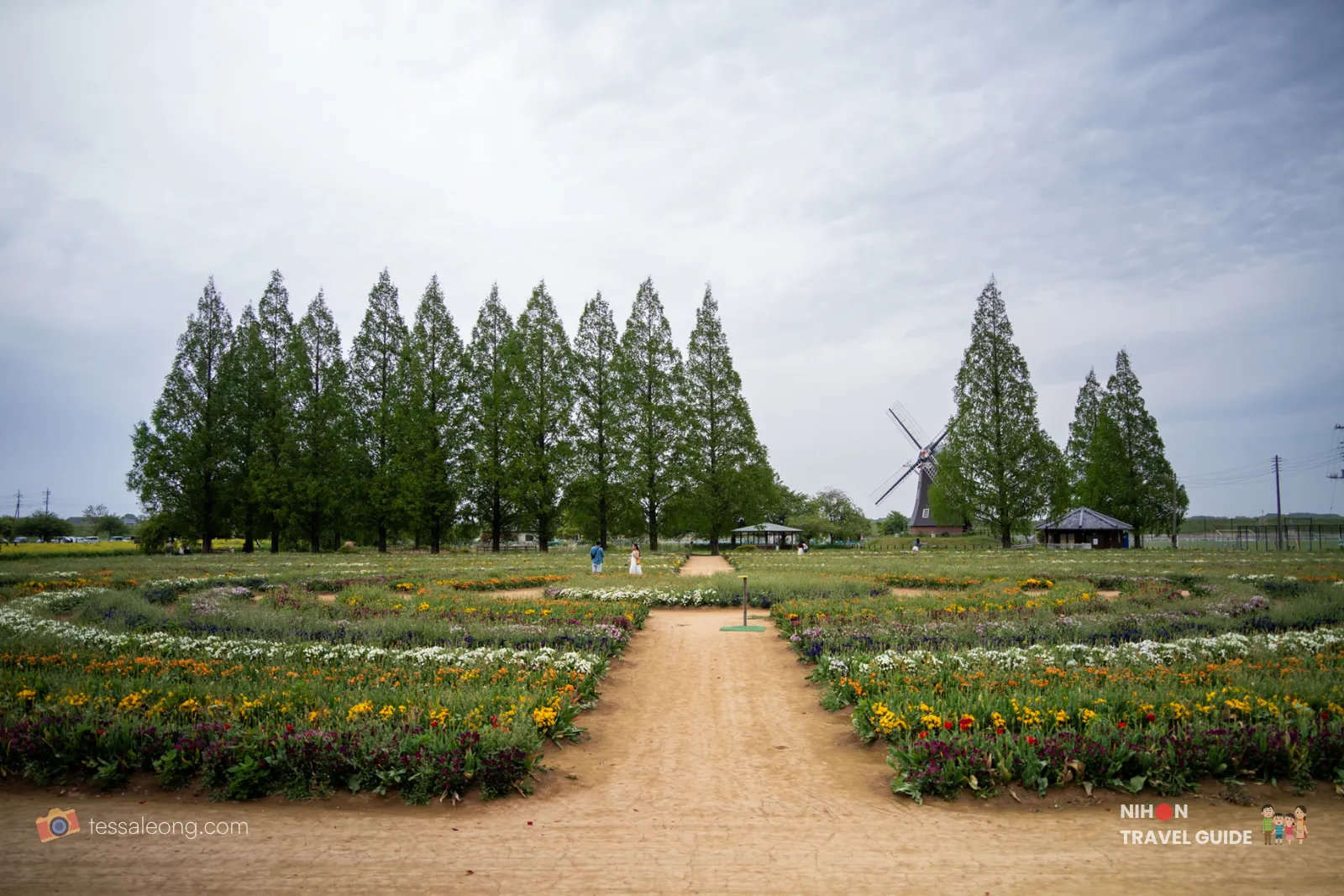 Spacious garden with a central dirt path, bordered by colorful flower beds, tall trees, and a windmill in the distance.