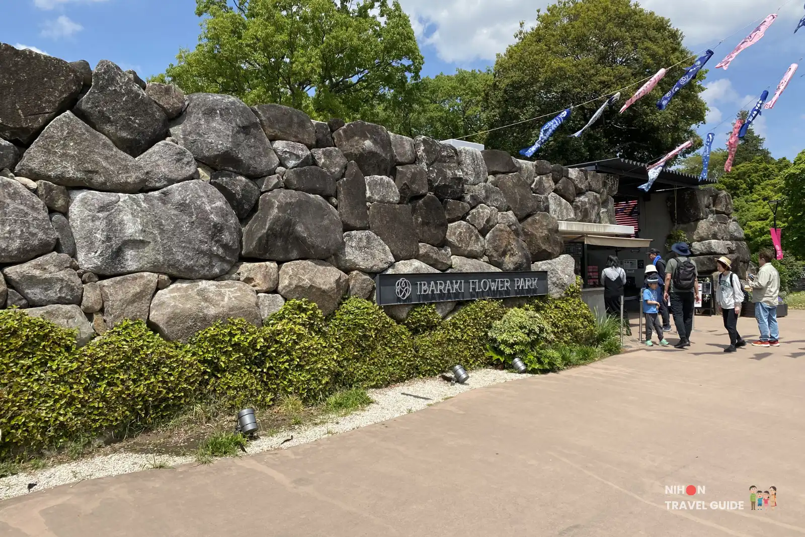 Stone rock wall entrance to Ibaraki Flower Park with a ticket booth and visitors nearby under colorful banners.