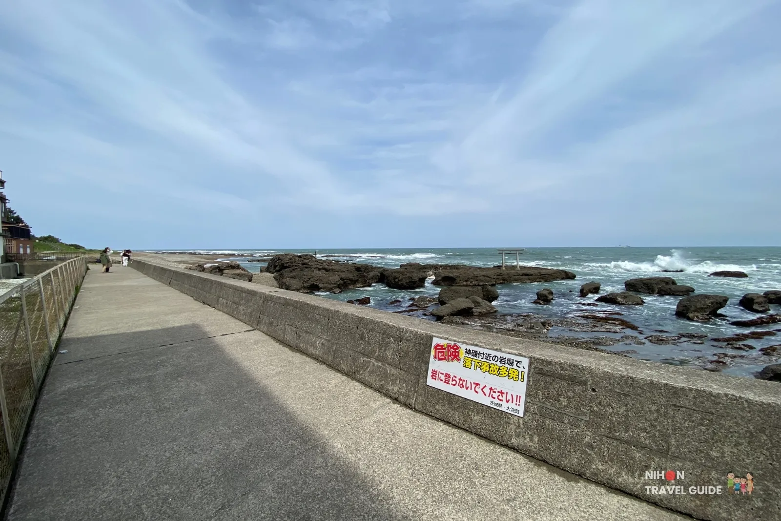 Concrete seawall path along the coast with a warning sign and view of the sea torii at Oarai Isosaki Shrine.