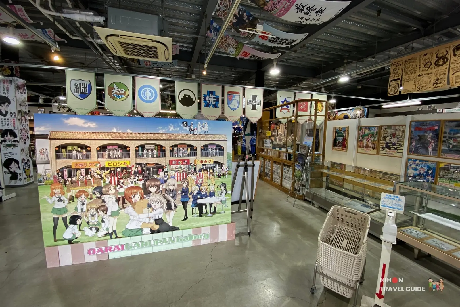 Indoor anime gallery or store displaying a large colorful illustration board featuring many characters, with banners hanging from the ceiling and glass display cases along the walls in the background.