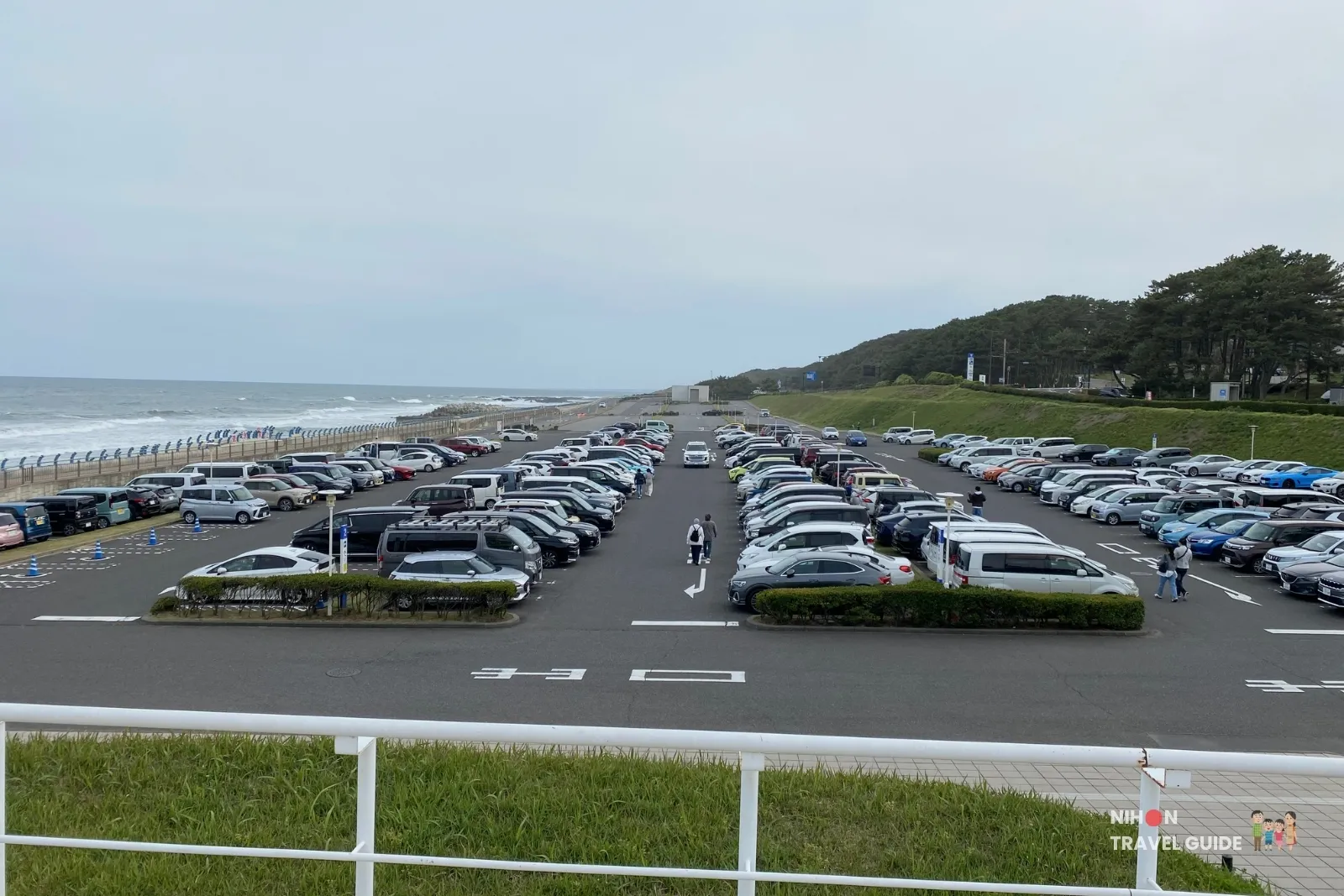 Large free parking lot at Aqua World Ibaraki Oarai Aquarium overlooking the Pacific Ocean, with rows of cars and the coastline in the background on a cloudy day.