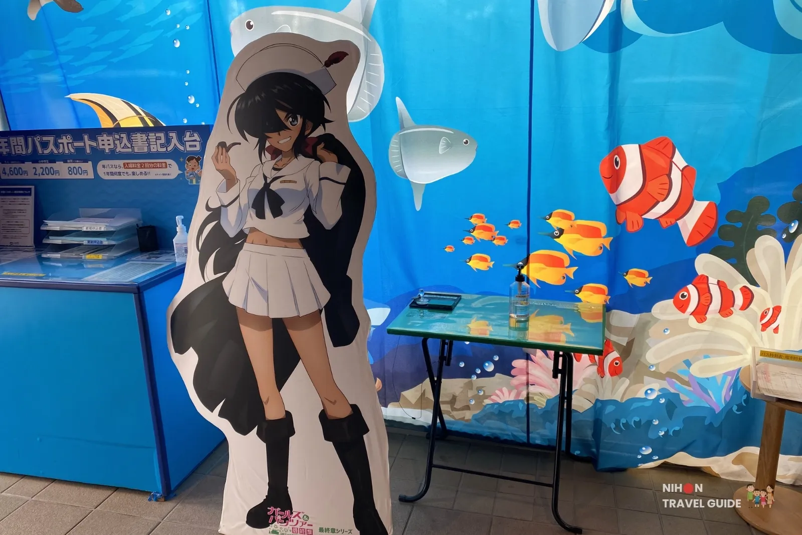 Girls und Panzer collaboration character standee at Aqua World Oarai Aquarium for the stamp rally event, with annual passport application desk and underwater-themed backdrop.