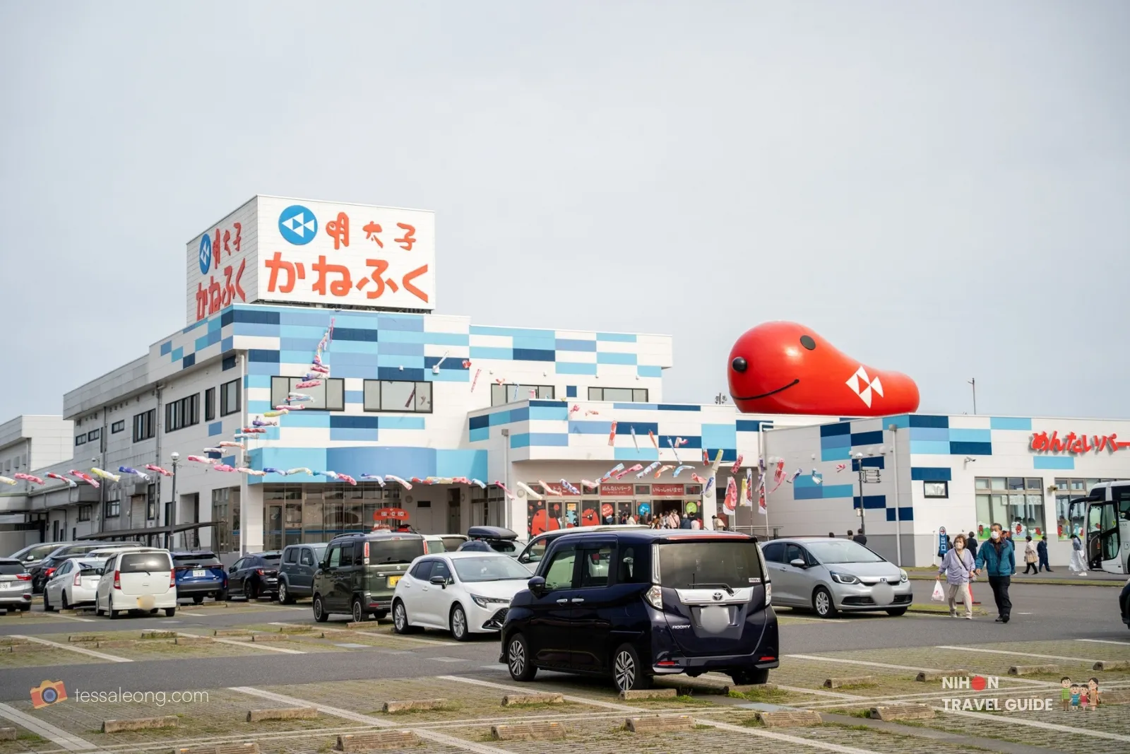 Exterior of Mentai Park Oarai (Kanefuku mentaiko factory) in Ibaraki, Japan, showing the blue-and-white checkered building, large red smiling mentaiko mascot balloon, parking lot with cars, and visitors under a cloudy sky.
