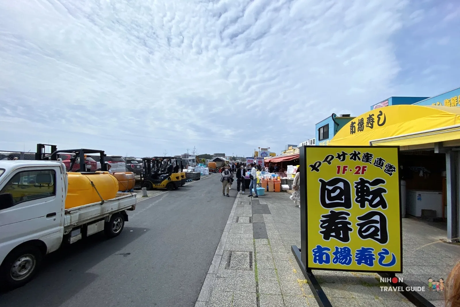 Yellow conveyor-belt sushi sign and bustling outdoor area at Nakaminato Fish Market in Hitachinaka, Ibaraki, with forklifts, visitors, and market stalls under a cloudy sky.