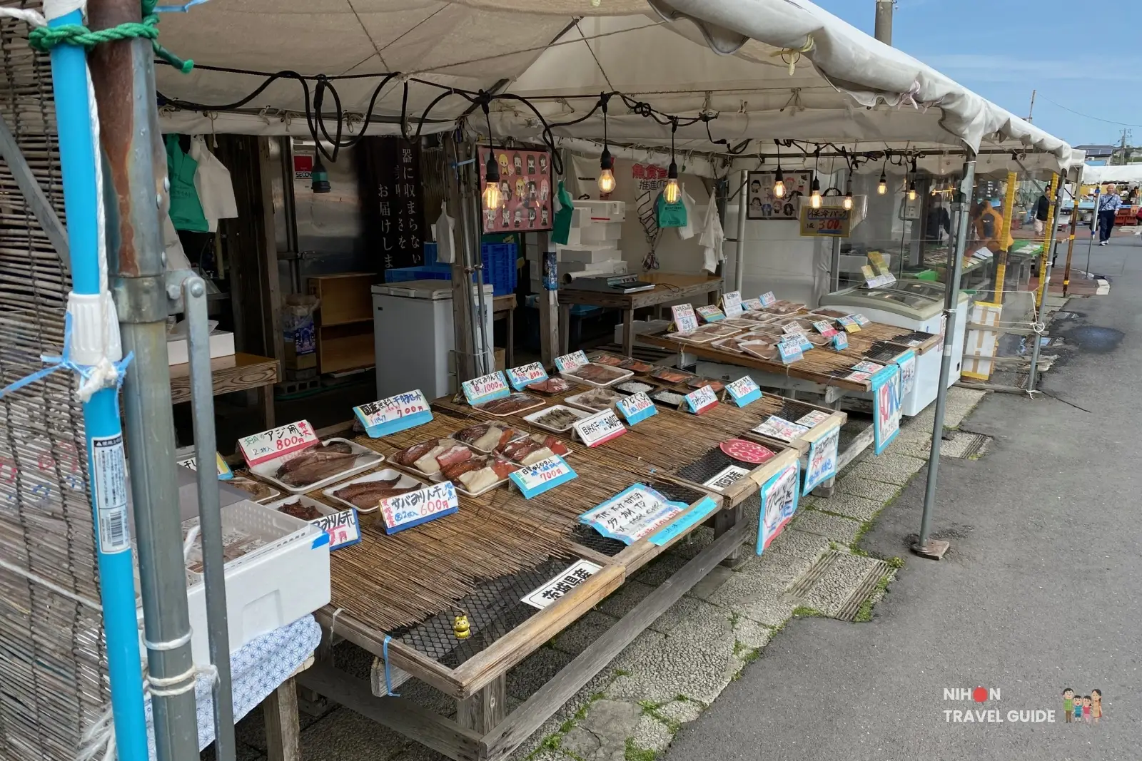 Dried fish (himono) stall at Nakaminato Fish Market displaying aji, saba, and other seafood with price signs on bamboo mats.
