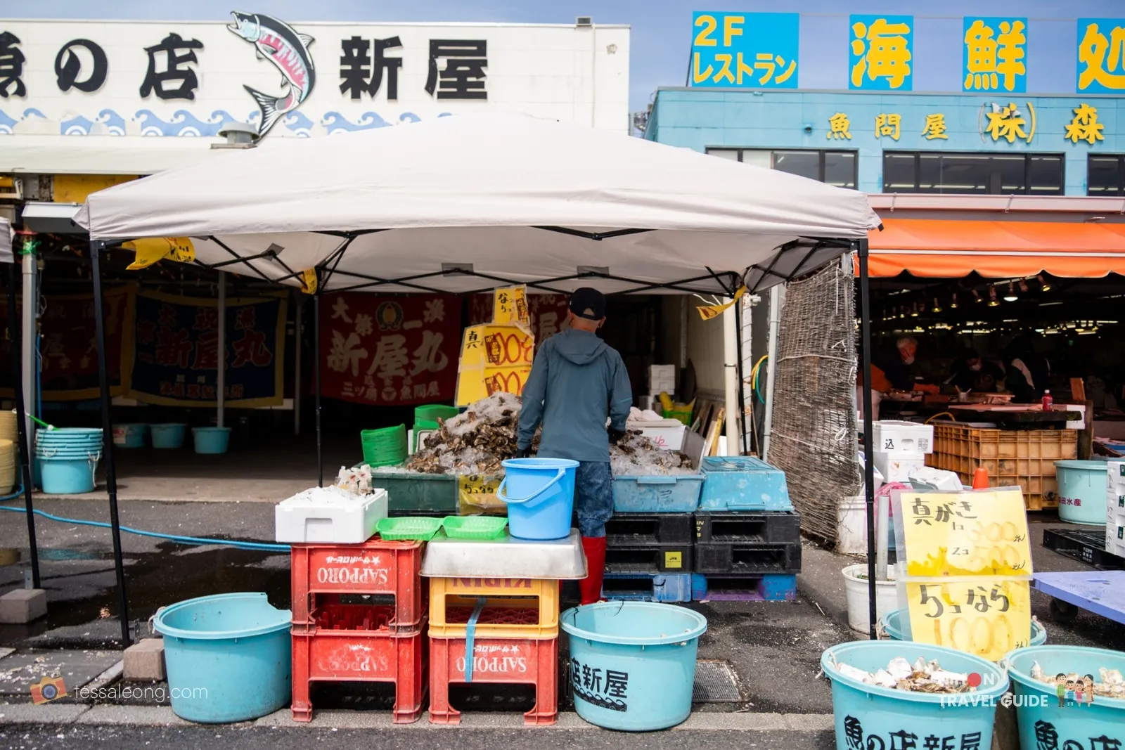 Fresh oyster stall with vendor working under a tent at Nakaminato Fish Market, surrounded by crates, buckets, and price signs in Ibaraki.