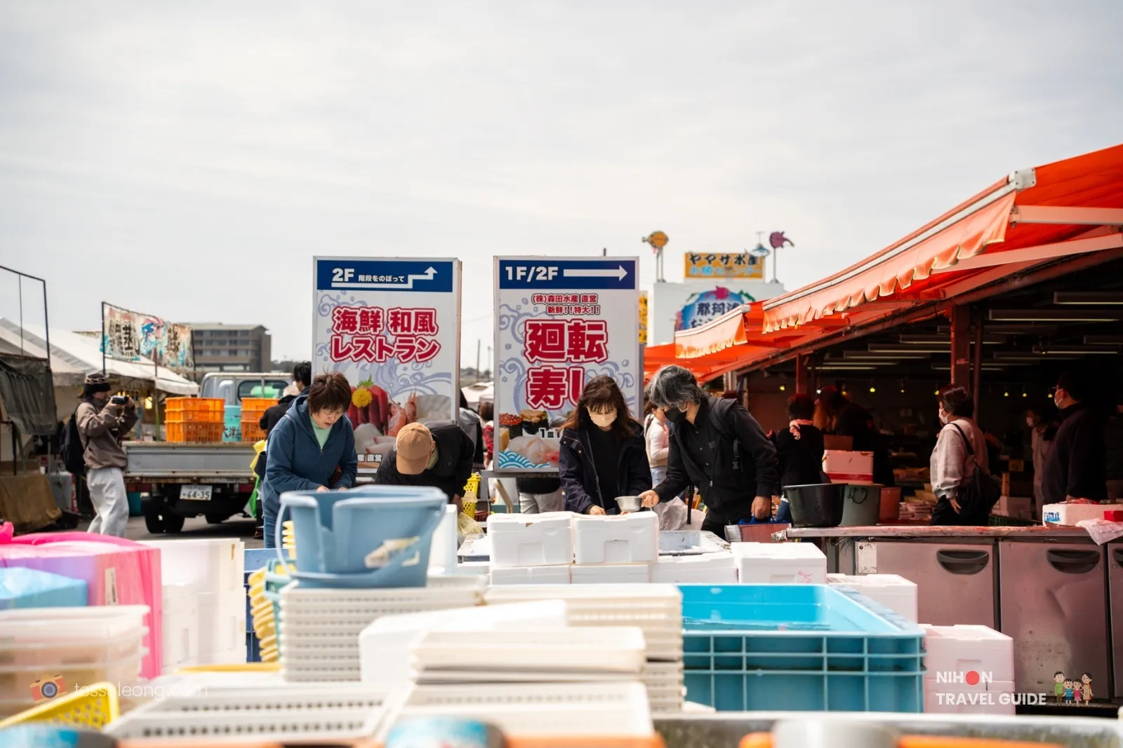 Outdoor seafood market with vendors and shoppers under orange canopies; stacked crates and coolers in the foreground, signs in Japanese directing to upstairs restaurants and markets.