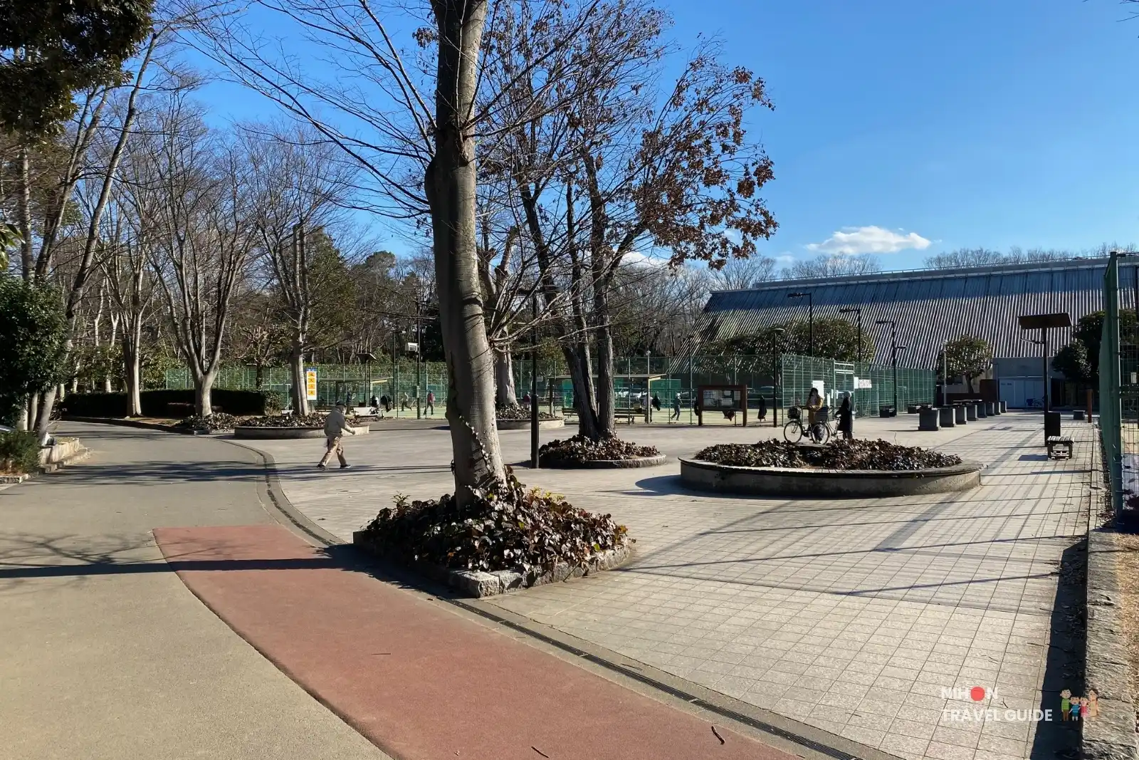 Tennis courts and paved plaza area at Doho Park Tsukuba in winter with people and the arena building in the background.