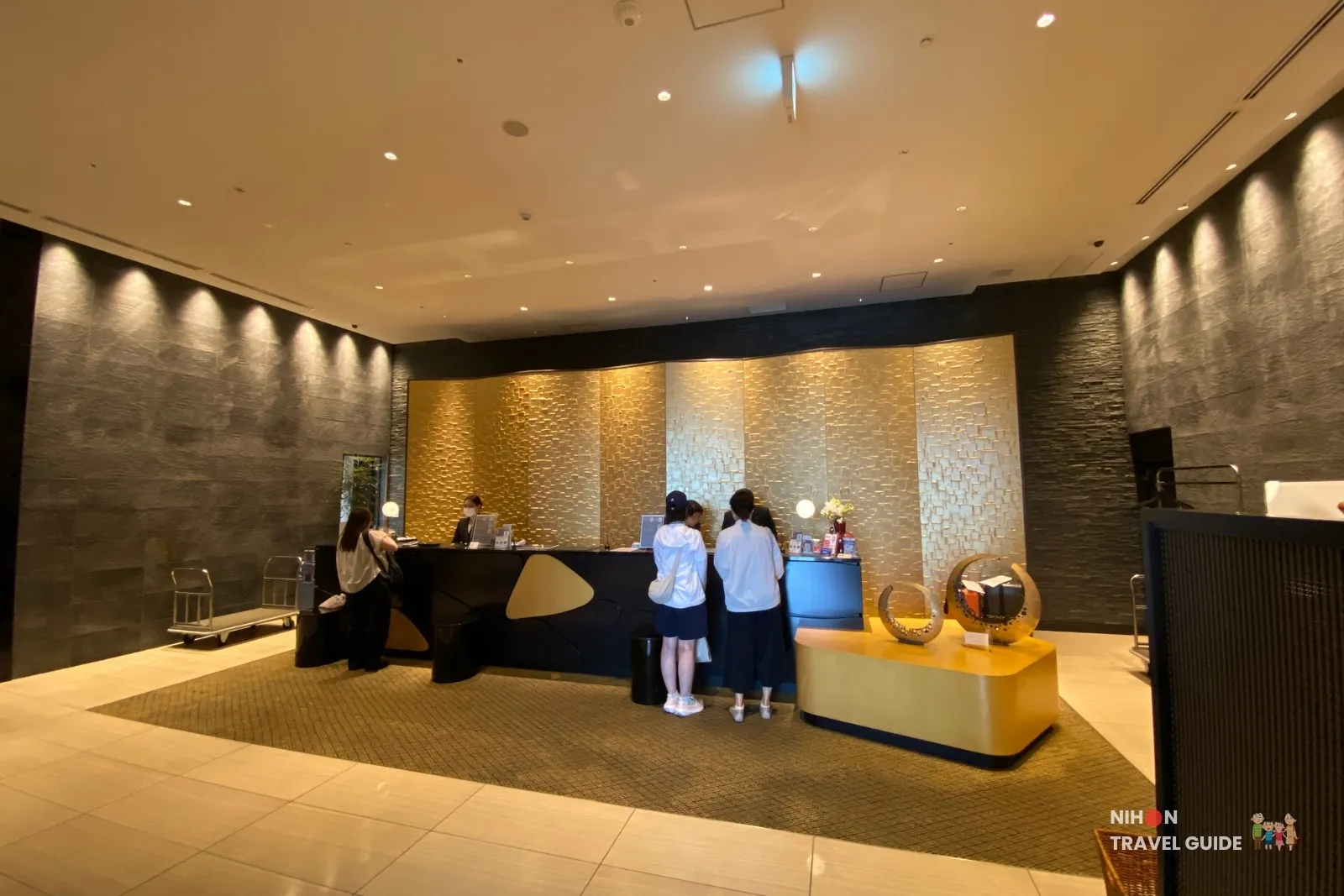 Modern check-in lobby of KOKO HOTEL Premier Kumamoto featuring a sleek black reception desk, gold textured wall, and contemporary design.