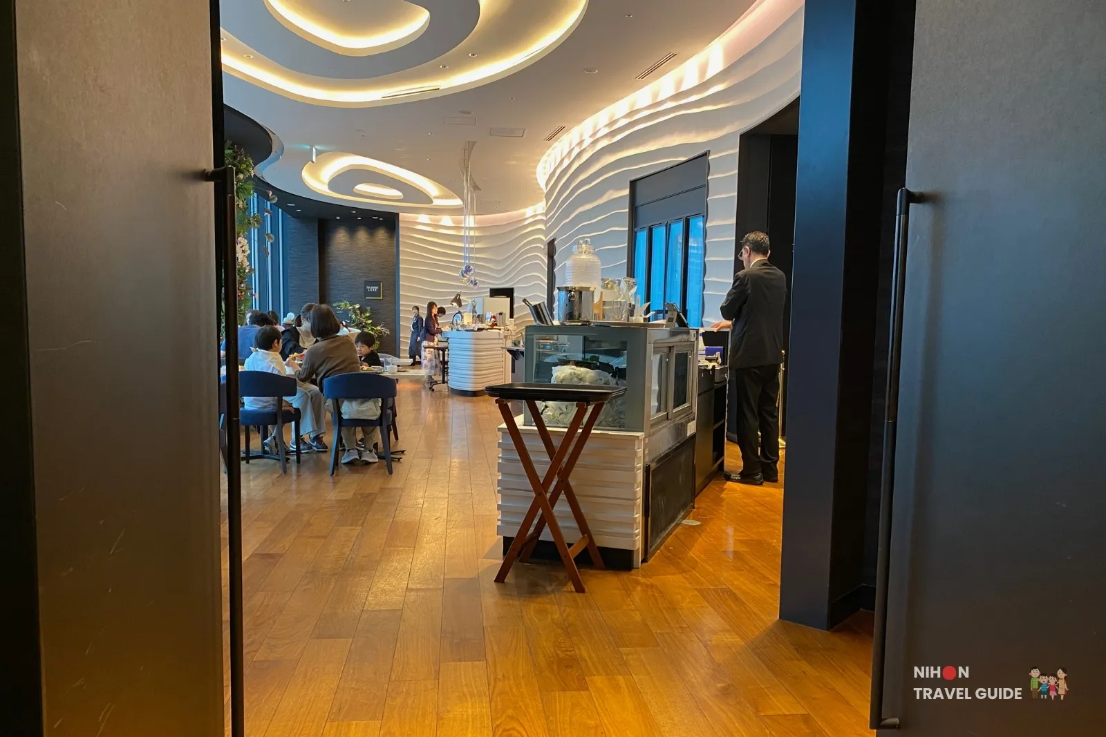 Interior of THE MOMENT restaurant at KOKO HOTEL Premier Kumamoto, featuring modern wave-patterned walls, wooden flooring, and guests dining with city views.