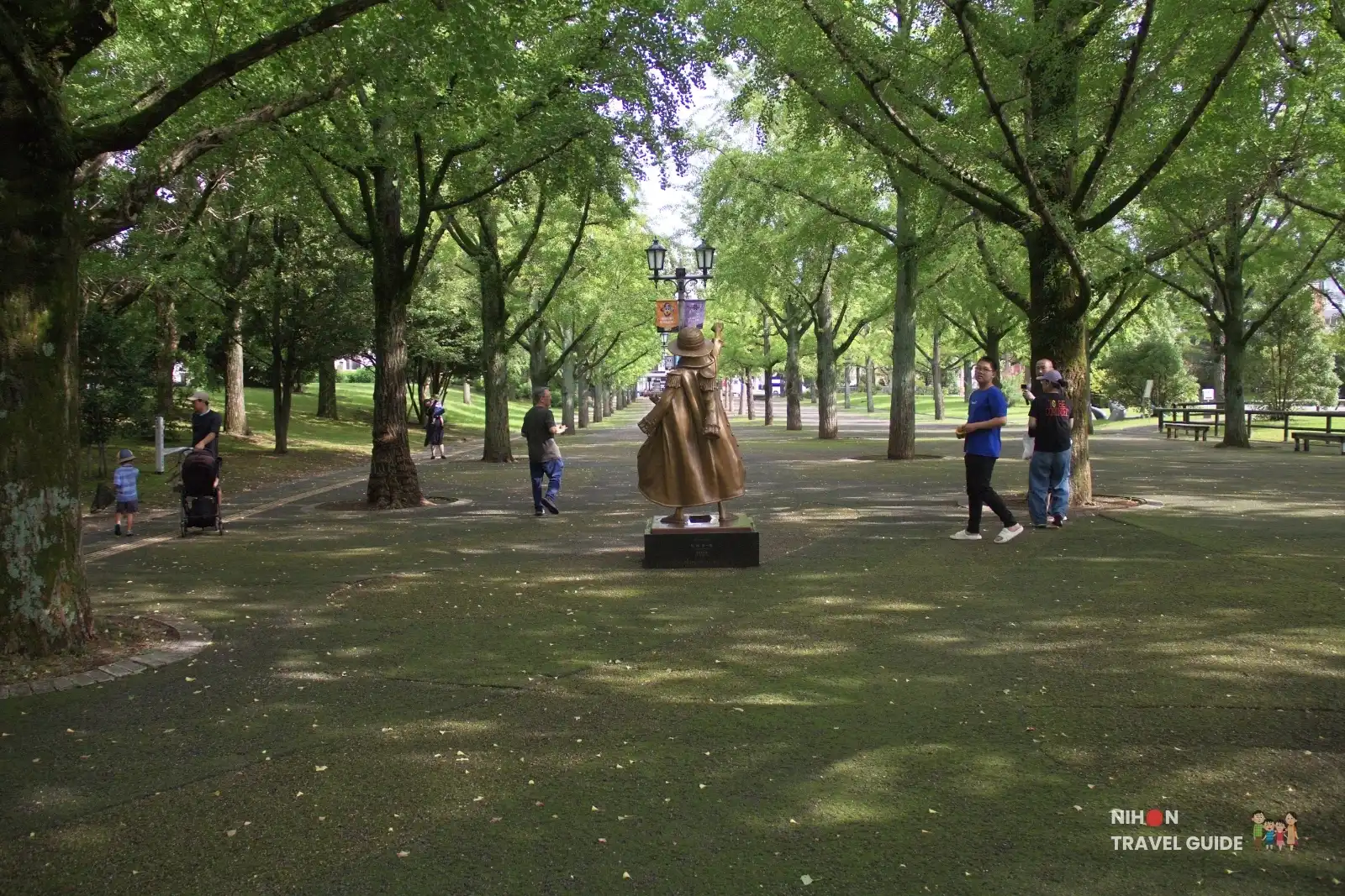 Rear view of the bronze Luffy statue from One Piece under the trees at Kumamoto Prefectural Government Office with visitors nearby.