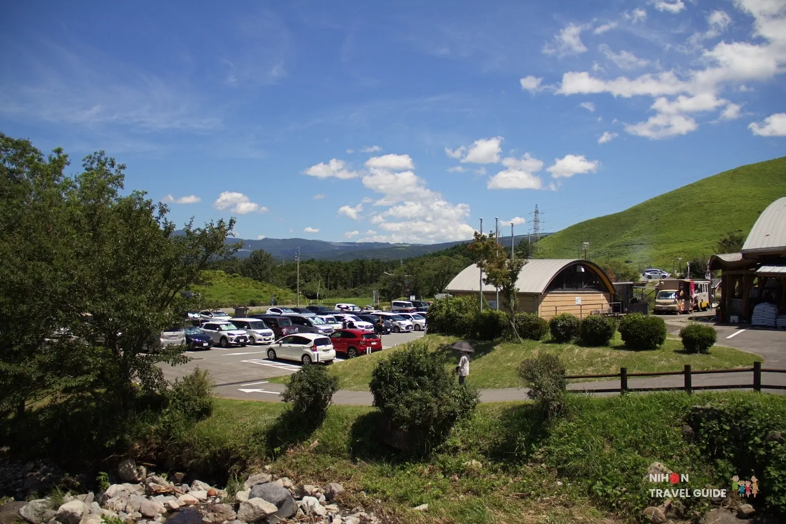 Parking lot and wooden buildings at Moe no Sato surrounded by green hills in the Aso region of Kumamoto.