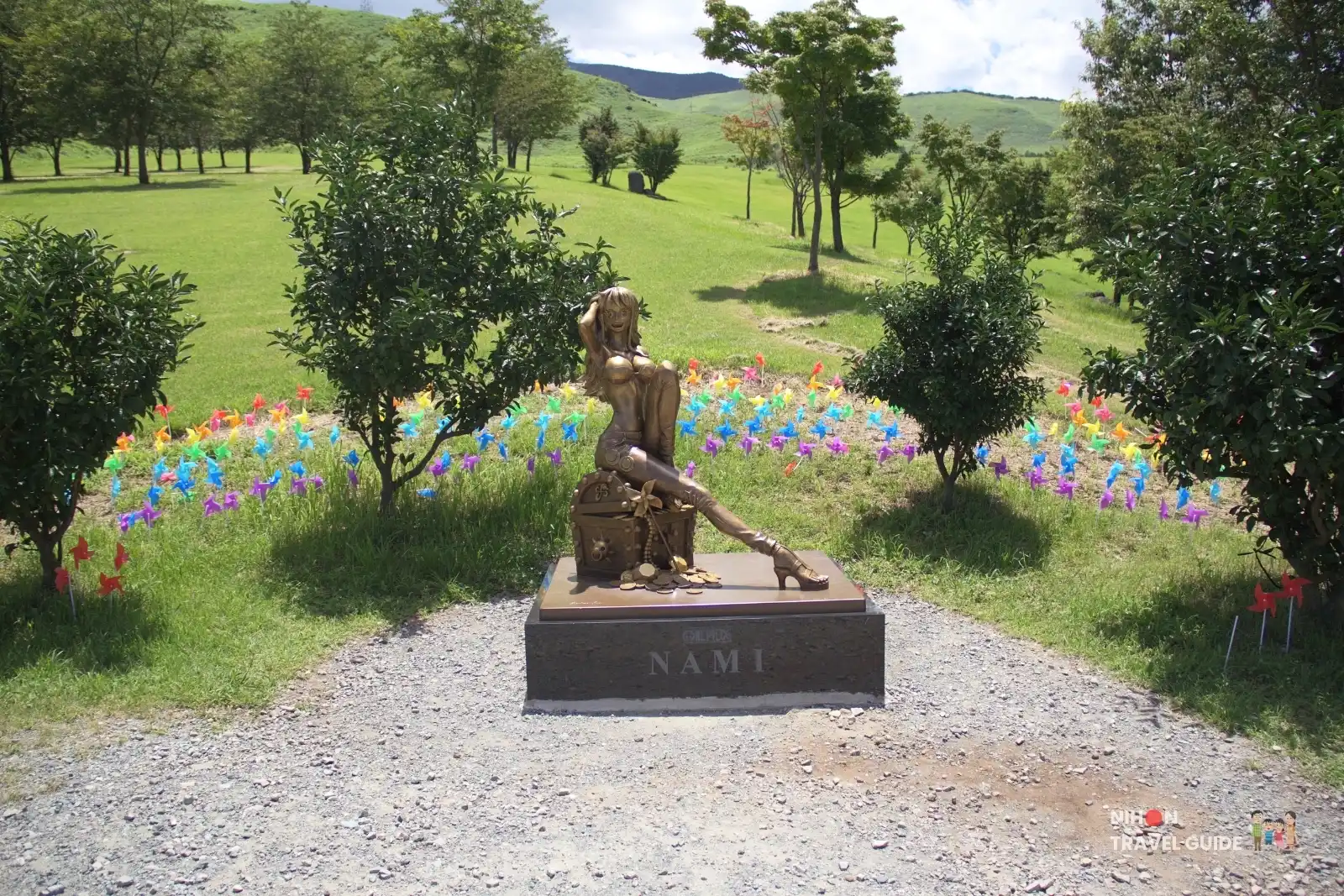 Wide view of the bronze Nami statue from One Piece sitting on a treasure chest surrounded by colourful pinwheels at Moe no Sato in Kumamoto.
