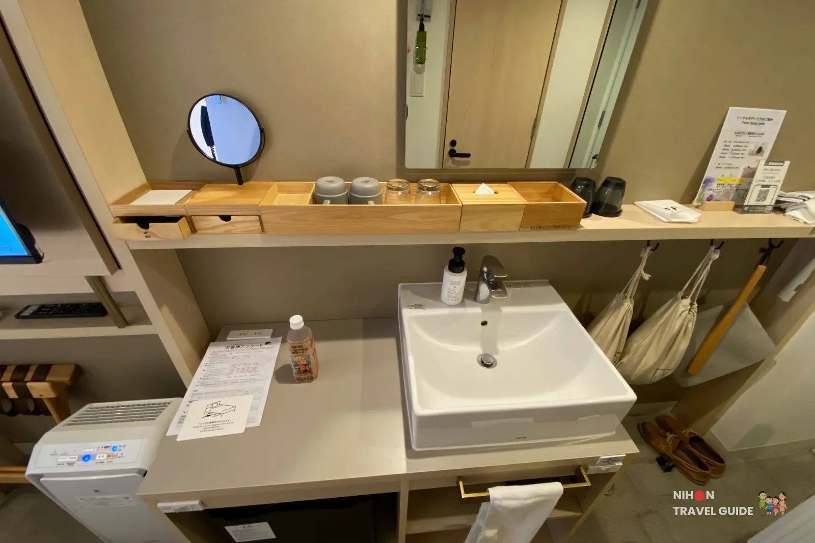 Open sink vanity area in the Single Room with Shower Booth at REF Kumamoto by Vessel Hotels, with wooden shelves, cups, toiletries, hand soap, air purifier and complimentary water bottle.