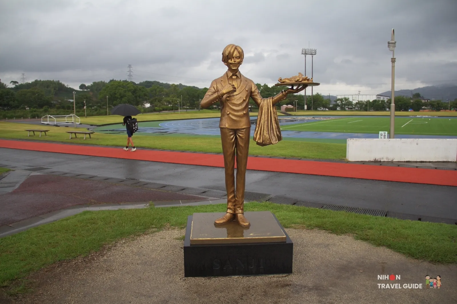 Full front view of the bronze Sanji statue from One Piece holding a food tray at Mashiki Town Sports Park in Kumamoto on a rainy day.